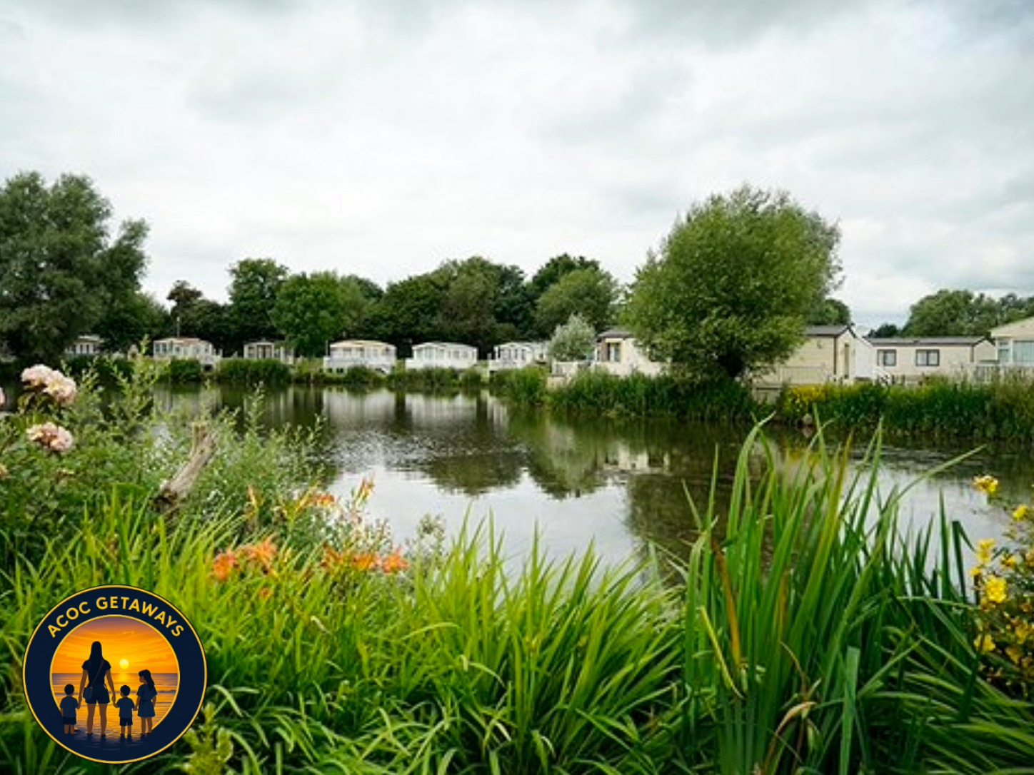 A peaceful scene of a small lake or pond surrounded by greenery, with several white houses with dark roofs in the background and lush trees, some flowering plants in the foreground, and cloudy sky overhead.