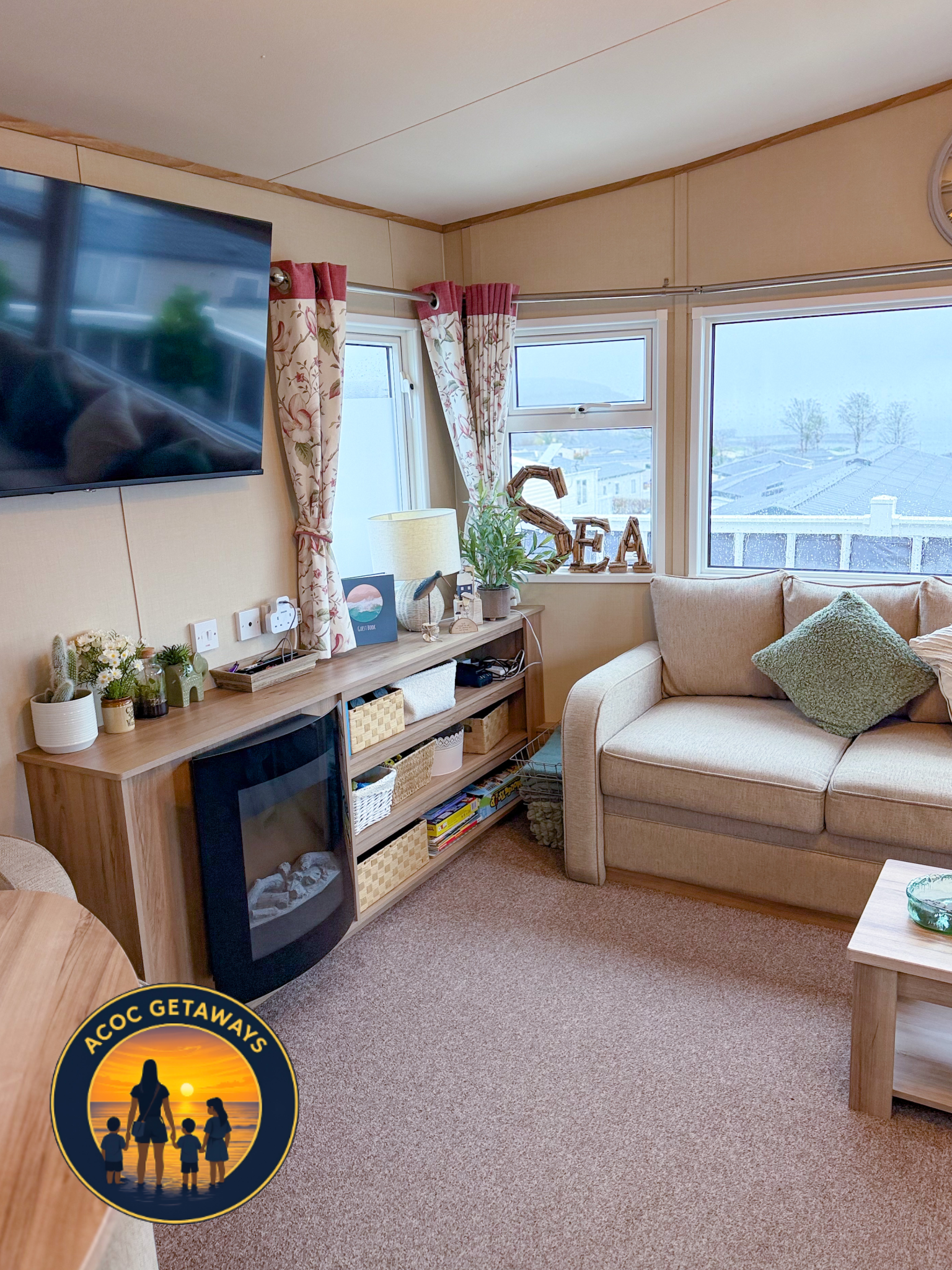 Living room with beige sofa, wooden coffee table, TV on the wall, window with floral curtains, decorative plants, and a sign spelling 'SEA' on the window ledge.