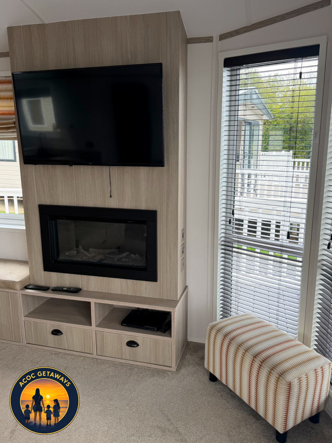 Living room with wall-mounted flat-screen TV, electric fireplace below, beige wooden entertainment unit, remote controls on the unit, striped upholstered ottoman, window with blinds showing outside view of houses and trees.