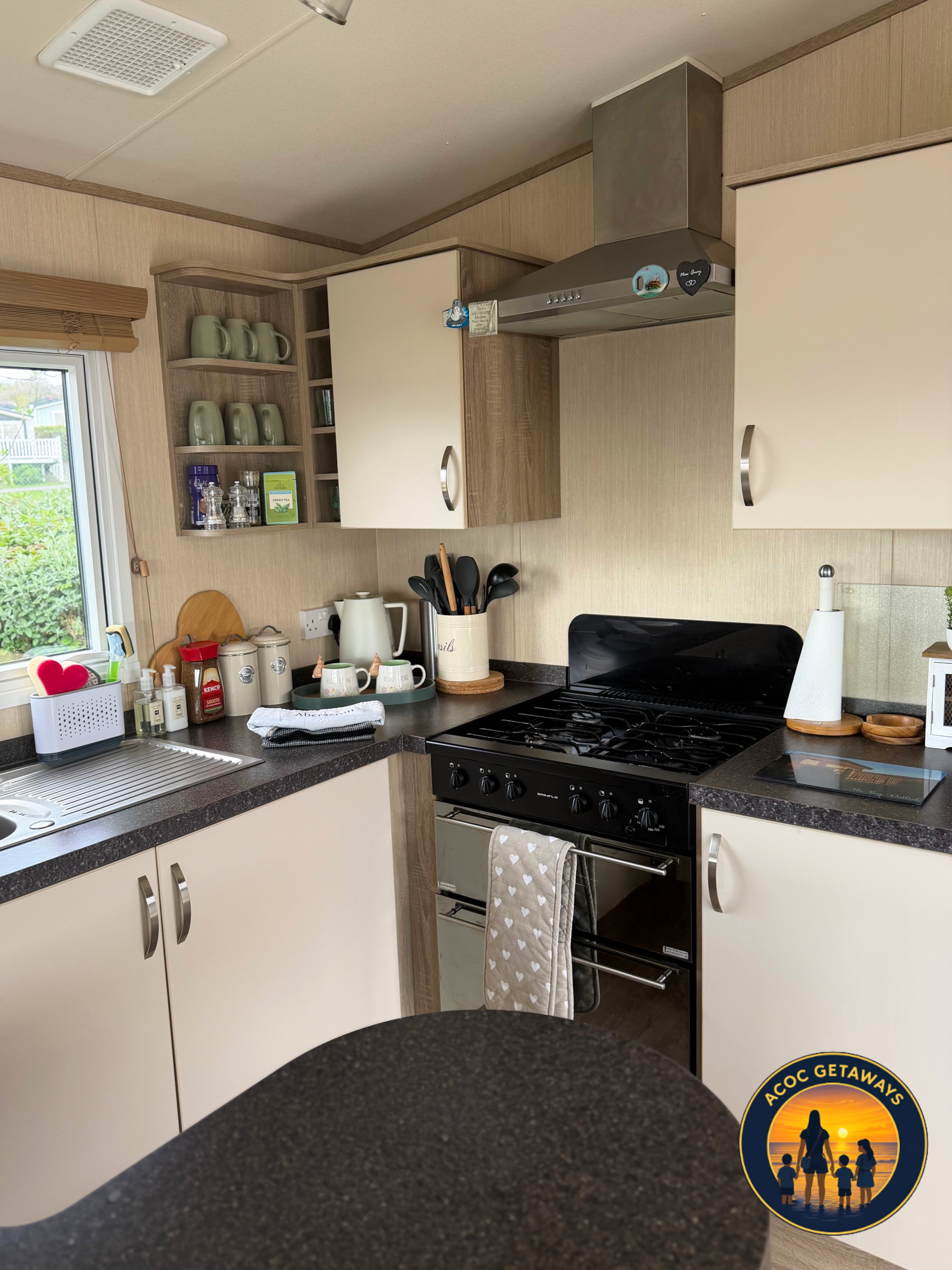 A kitchen with beige and wood cabinets, a black stove, a white kettle, and various kitchen items on the counters. There are open shelves with mugs and glassware and a window with a green outdoor view.