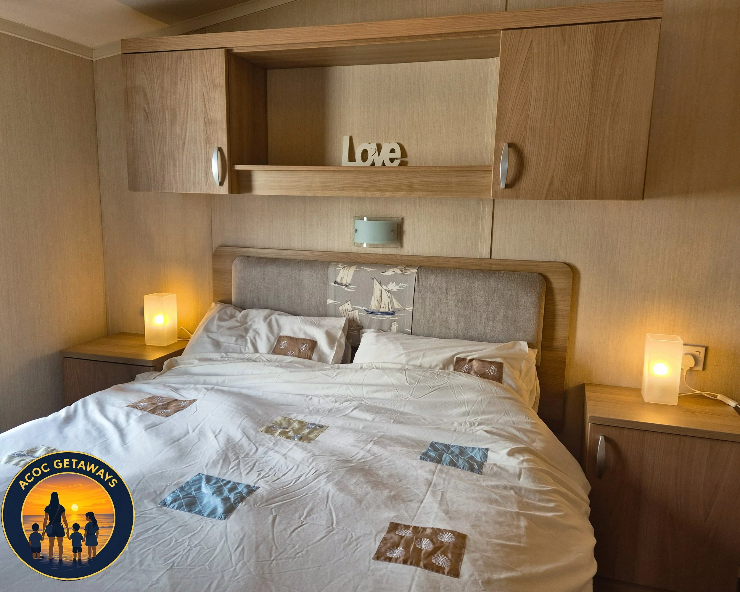 A neatly made bed with white bedding and decorative pillows, flanked by two wooden nightstands with modern lamps. There is a wooden headboard and a wall-mounted light above. A wooden shelf above the bed displays a decorative "Love" sign.
