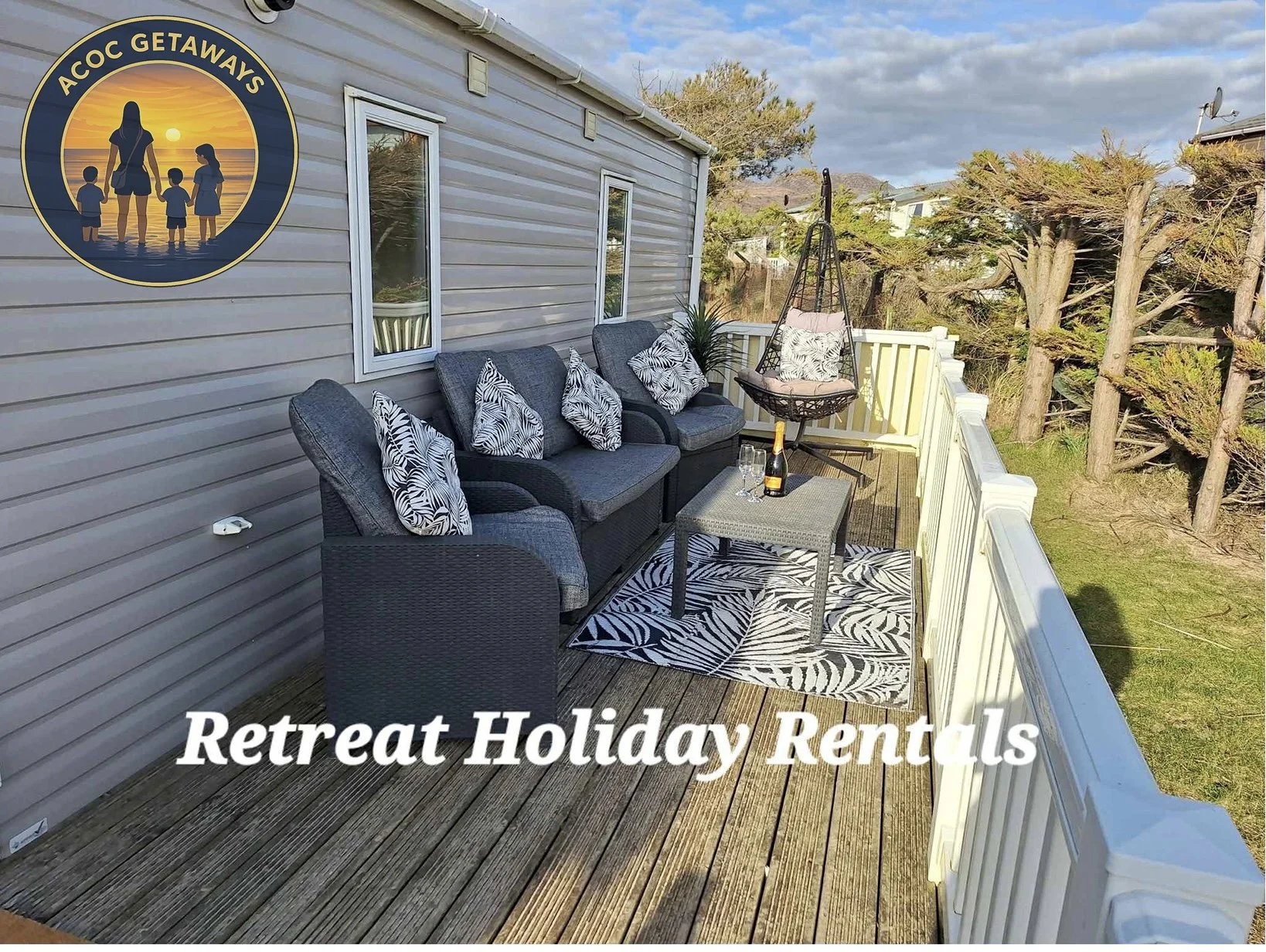 An outdoor patio with a black wicker sofa, matching armchair, a small table with a wine bottle and glasses, zebra print cushions, a hanging chair with cushions, an outdoor rug with a zebra pattern, and a white railing, overlooking trees and a partly 