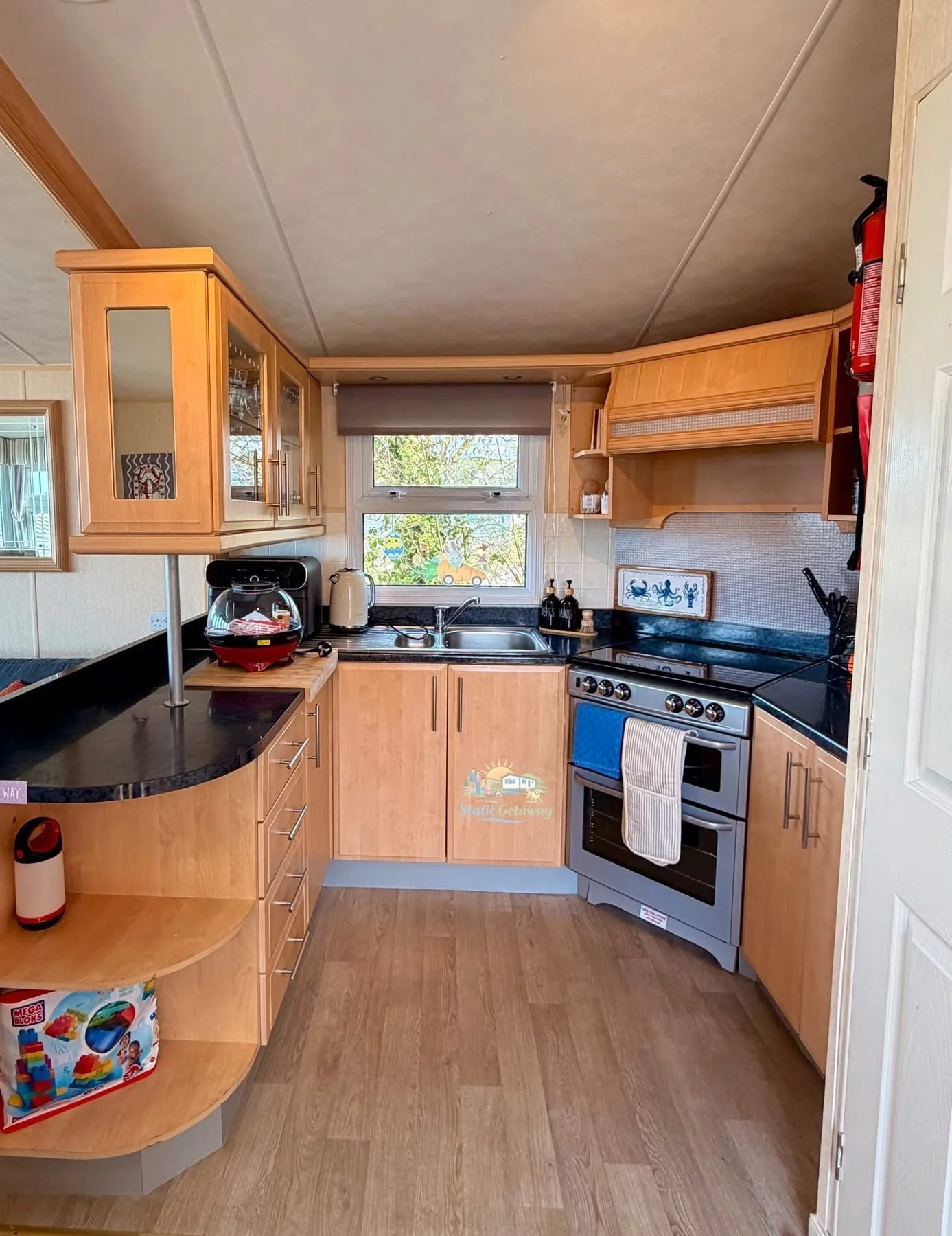 Kitchen with wooden cabinets, black countertops, oven, small window, and various appliances including a coffee maker and electric kettle. There are colorful stickers on the window, a framed picture of crabs on the wall, and a fire extinguisher mounte
