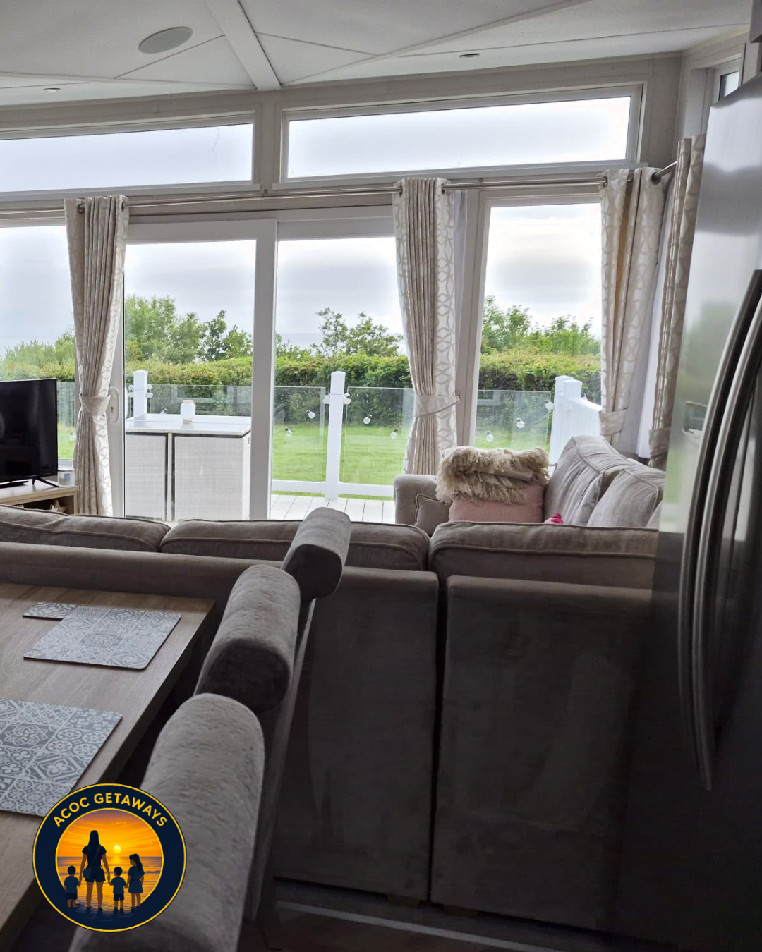 Living room with large windows and sliding glass door leading to a green outdoor area with trees; beige sectional sofa with cushions and throws; dining table with placemats; partial view of a TV and side table; curtains tied back.