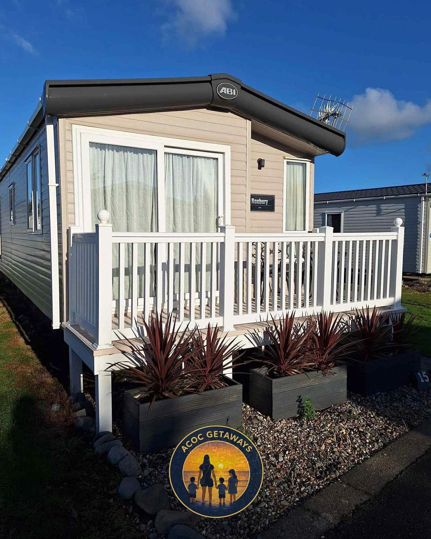 A small home with a wooden deck and white railing, beige siding, glass doors with curtains, a black awning with 'ABI' logo, plant boxes with reddish plants, and a sign that reads 'Roxbury' on the house. There is a charity badge at the bottom with a silhouette of a woman and children, reading 'ACOC GETAWAYS' with a sunset background.