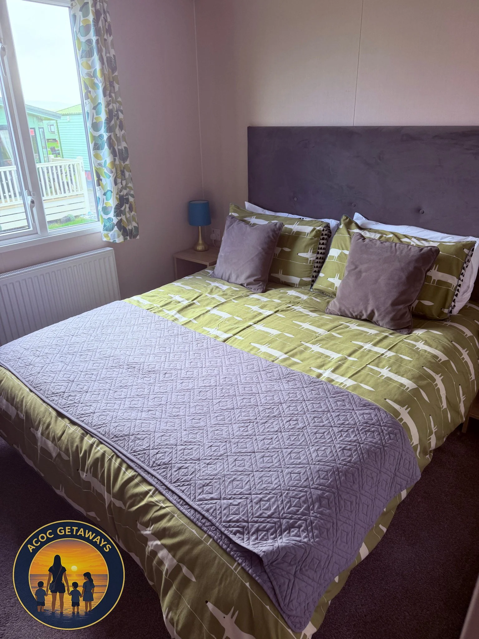 A neatly made bed with green and white patterned bedding, two beige pillows, and two brown pillows. There is a small bedside table with a blue lamp next to the bed. A window with colorful floral curtains shows a view of neighboring houses and a patio