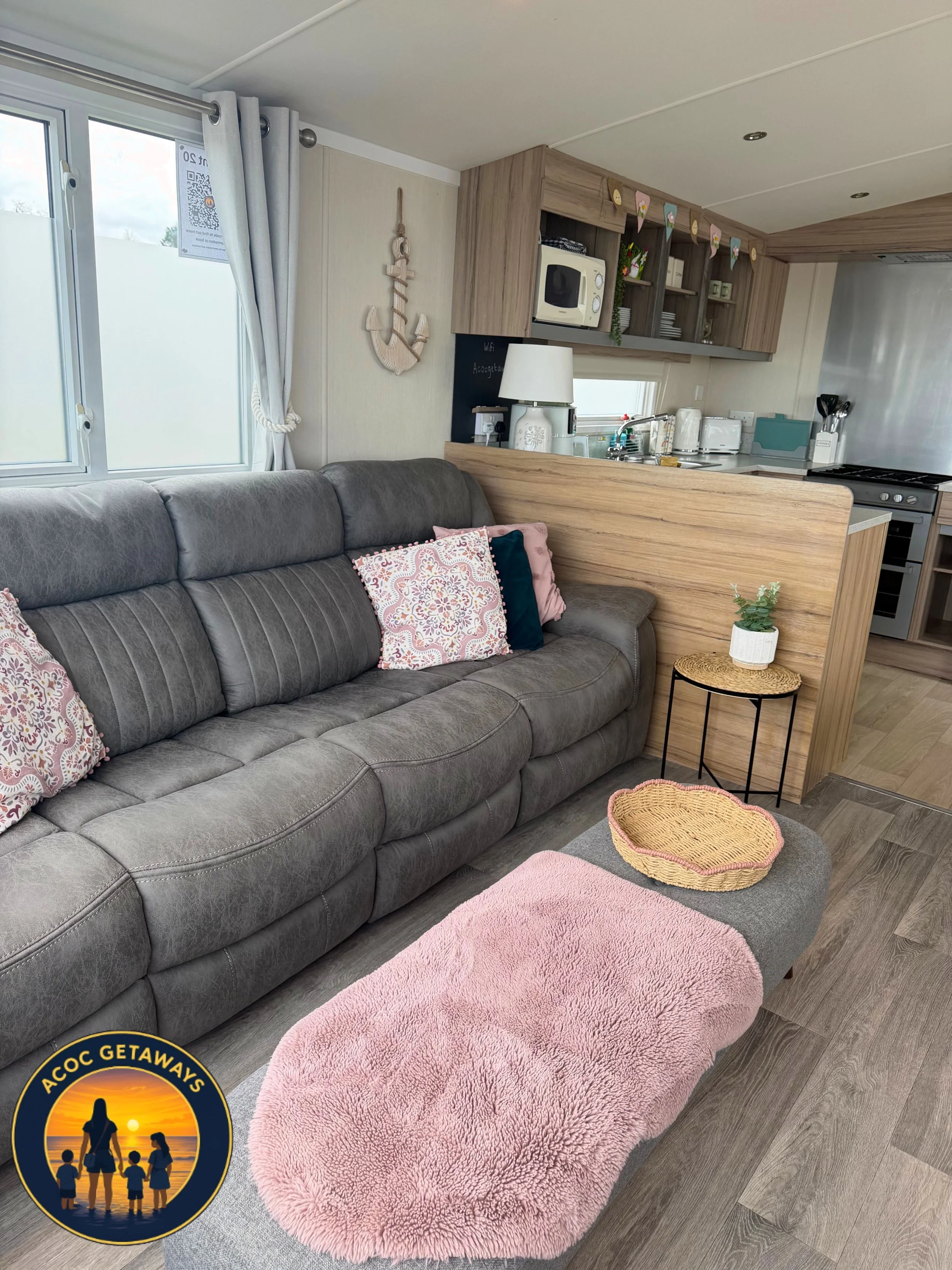 A cozy living room with a gray sofa adorned with pink and patterned throw pillows, a pink shaggy oval rug in front, a small round wicker side table with a plant, a kitchen area with wooden cabinets, a small window, and various appliances, and a decor