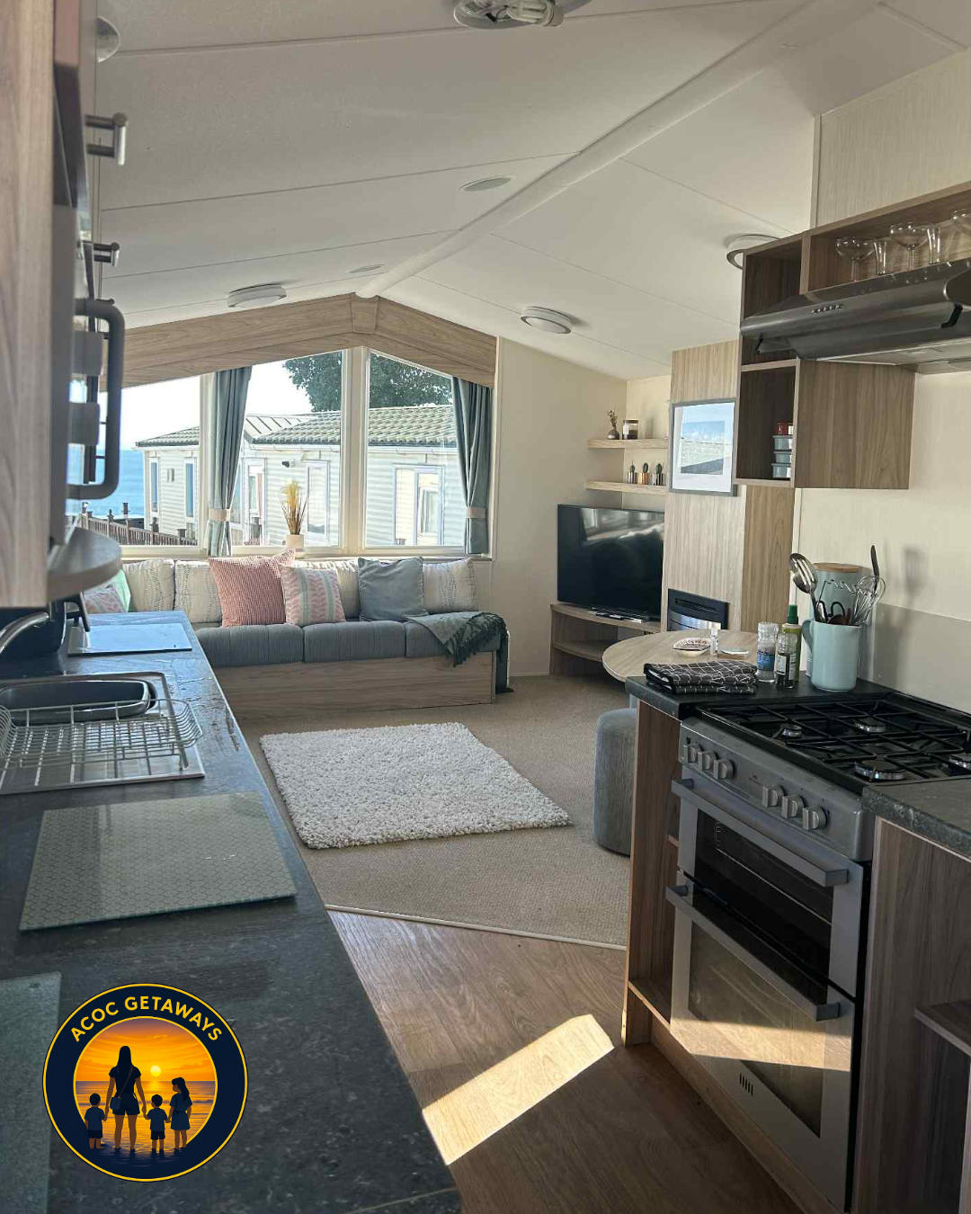 Interior of a cozy cabin or tiny home with a kitchen area on the right, a small dining table, and a living room with a window seat and TV on the far wall.