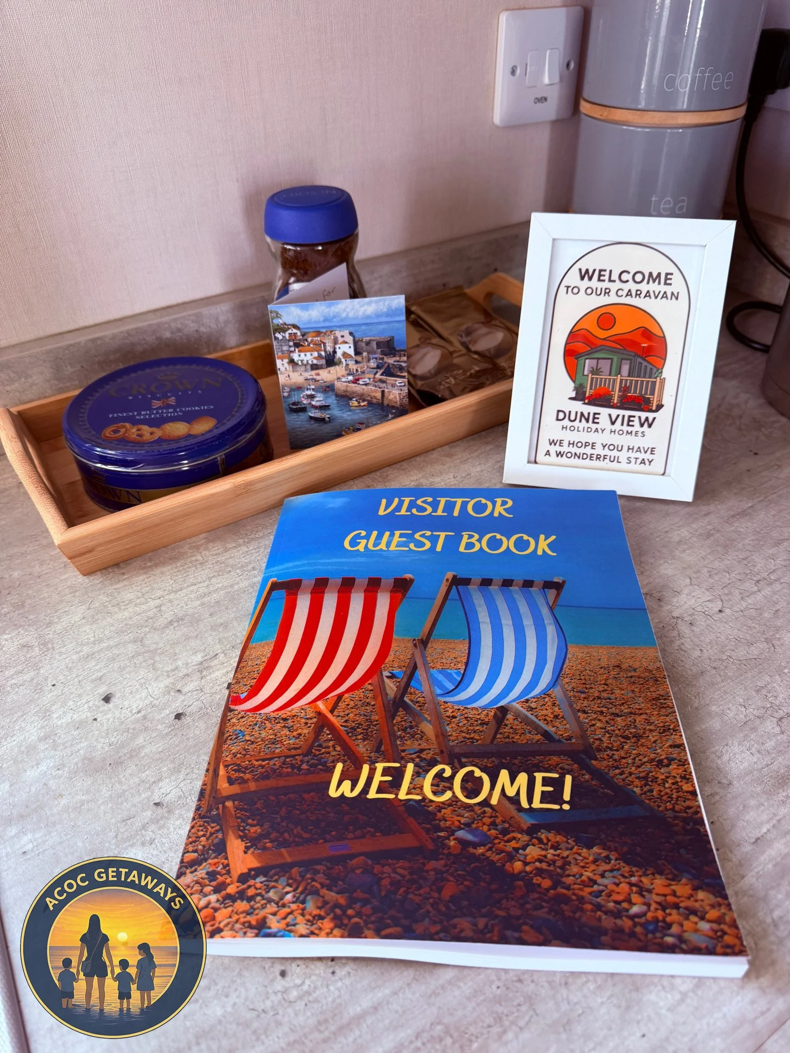 Visitor guest book titled "Welcome!" with an image of two beach chairs on a sandy beach. There is a welcome sign for Dune View holiday homes, a tray with treats, a framed photo, and a jar of cookies on a countertop.
