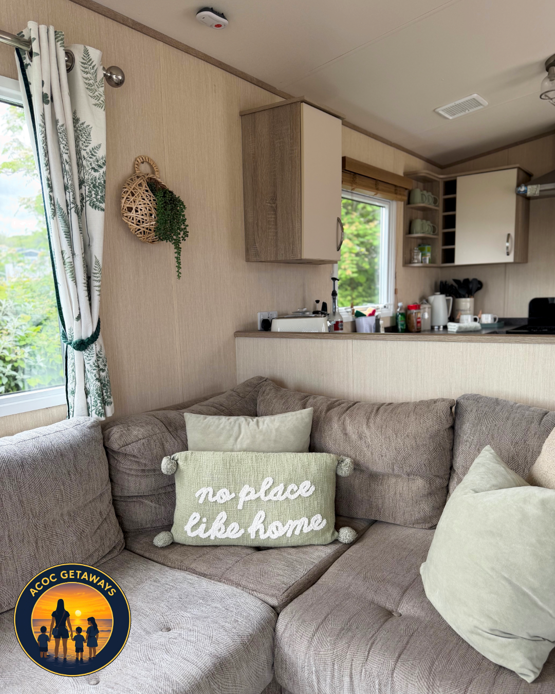 A cozy living room with a beige sectional sofa, decorated with pillows including a green pillow with white text saying 'no place like home,' in front of a window with patterned curtains. The open kitchen area is visible with cabinets, a countertop fi