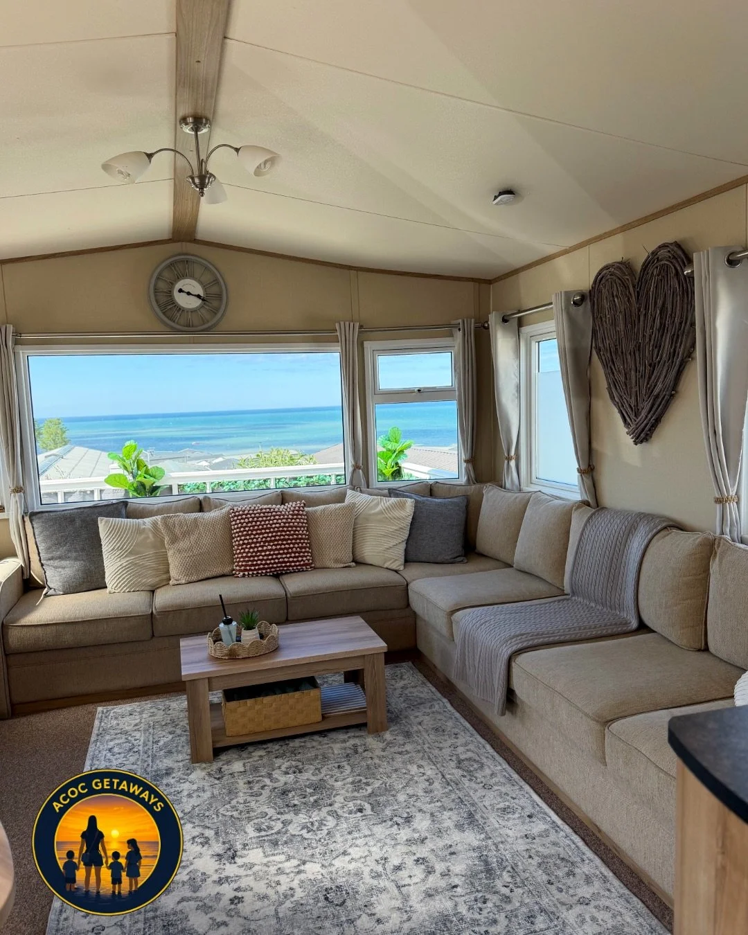 Cozy living room with large windows showing a view of the ocean, a sectional sofa with multiple pillows, a wooden coffee table, a decorative heart-shaped piece on the wall, and a ceiling fan with light fixtures.