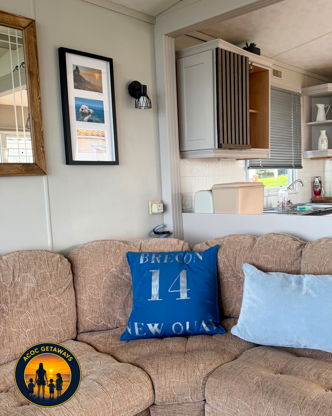 Living room with beige patterned couch, blue 'Brecon 14' pillow, light blue pillow, photo frame of scenic views and a dog, wall light, and view of kitchen with white cabinets, window with blinds, and kitchen sink.