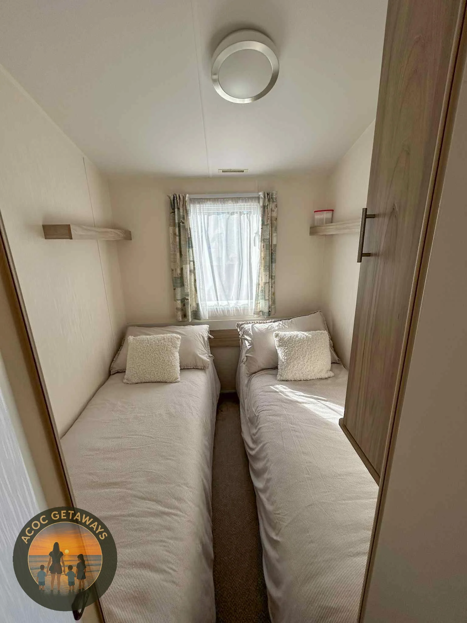 A small bedroom with two twin beds with white linens and fluffy pillows, facing each other with a window at the far end, curtains partly drawn, and a wooden wardrobe on the right side.