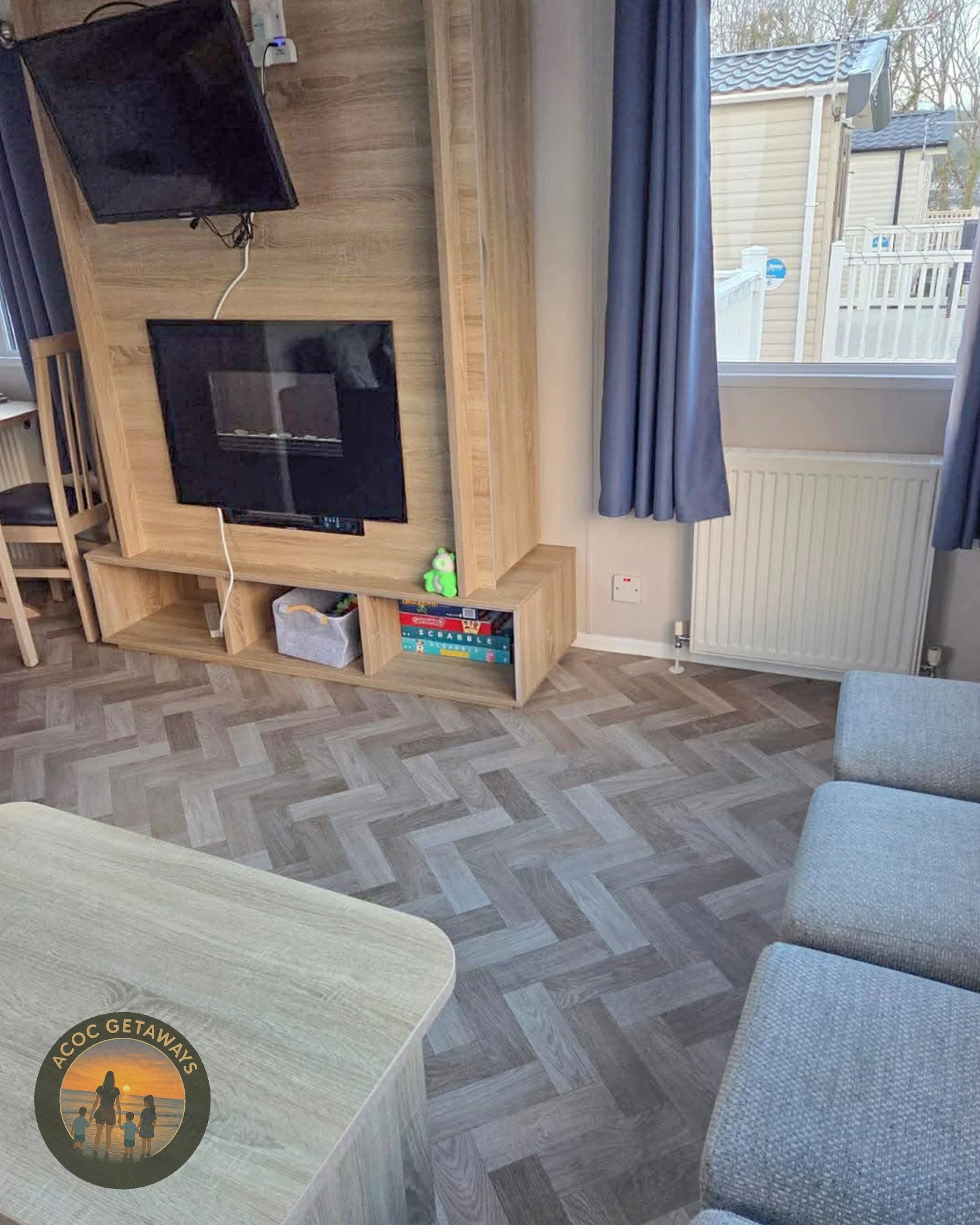 Living room with wooden entertainment unit featuring a wall-mounted TV, a fireplace below it, a small plush frog toy on the unit, and books. A window with blue curtains, gray chairs, and a light wood table are also visible.