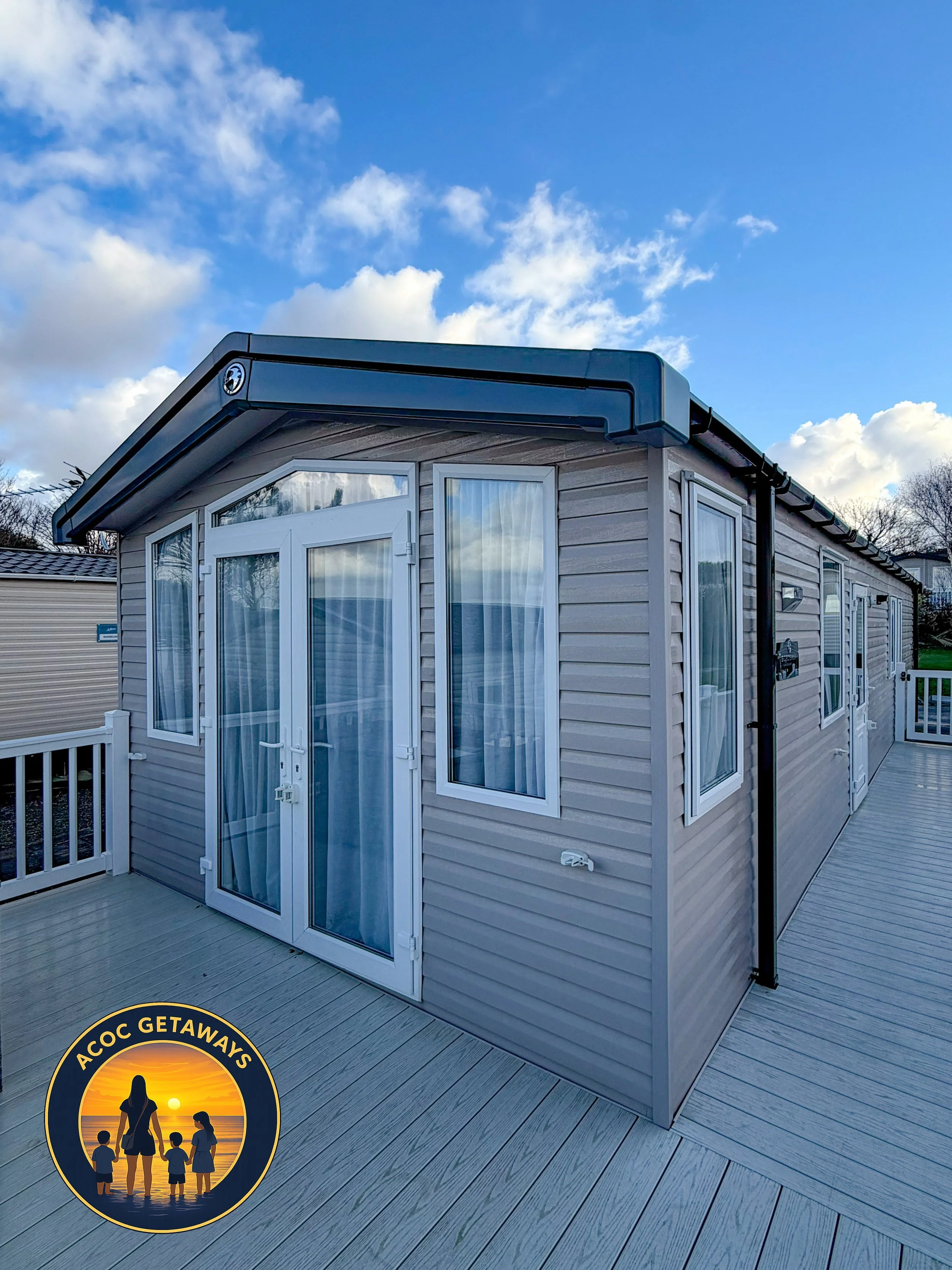 A tiny house with large windows and beige siding on a wooden deck, under a partly cloudy sky, with a logo showing a woman and children at sunset.