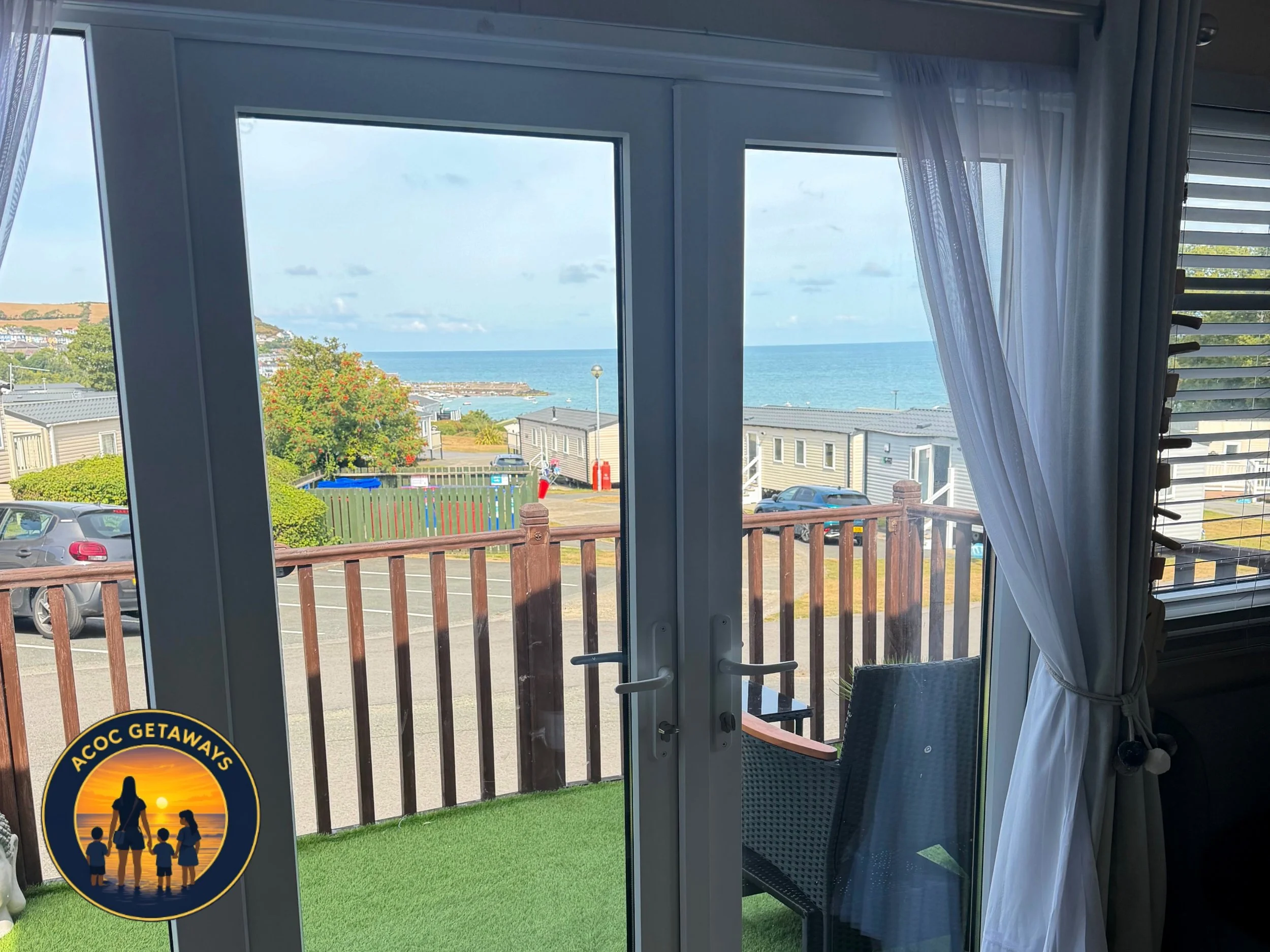 View of the ocean through a sliding glass door from inside a room with sheer curtains, a balcony with railing, parking lot, playground equipment, and houses in the background.