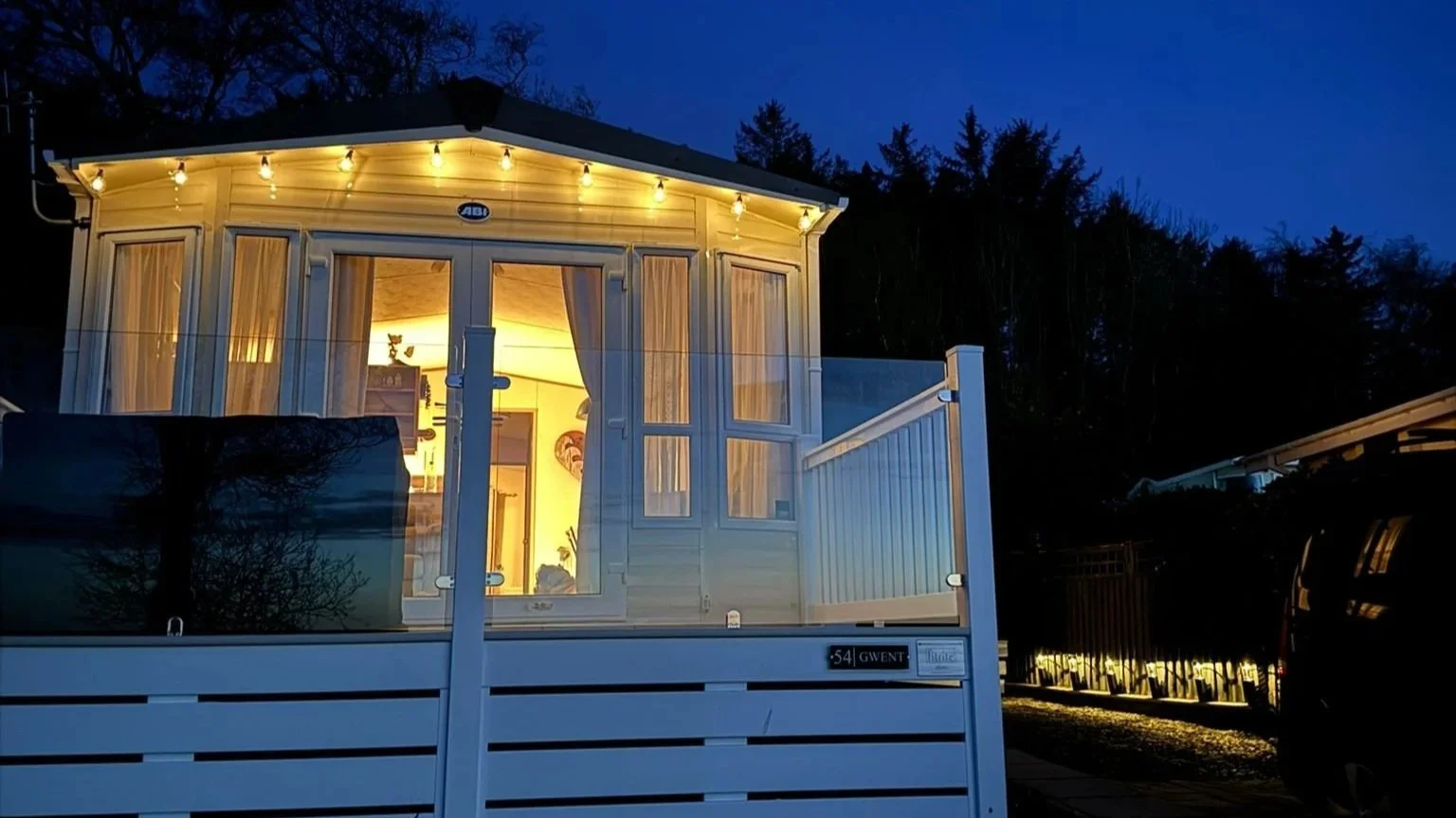 A small, cozy house with a porch, illuminated from inside at dusk with string lights hanging outside.