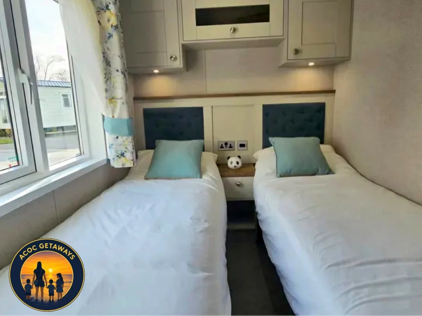 A bedroom with two single beds, each with a light blue pillow, white bedding, and a blue upholstered headboard. There is a window with floral curtains on the left side. Between the beds is a small nightstand with a panda-shaped lamp and electrical ou