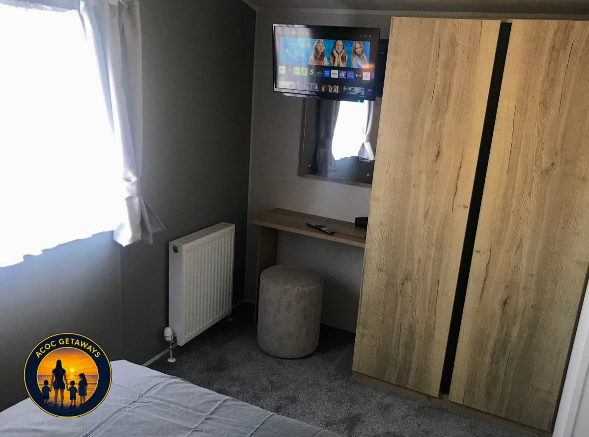 Small hotel room with a gray wall, a window with white curtains, a flat-screen TV mounted on the wall showing a TV guide, a mirror, a small desk with a remote control on it, a round beige stool, a wooden wardrobe, carpeted floor, and a bed with white