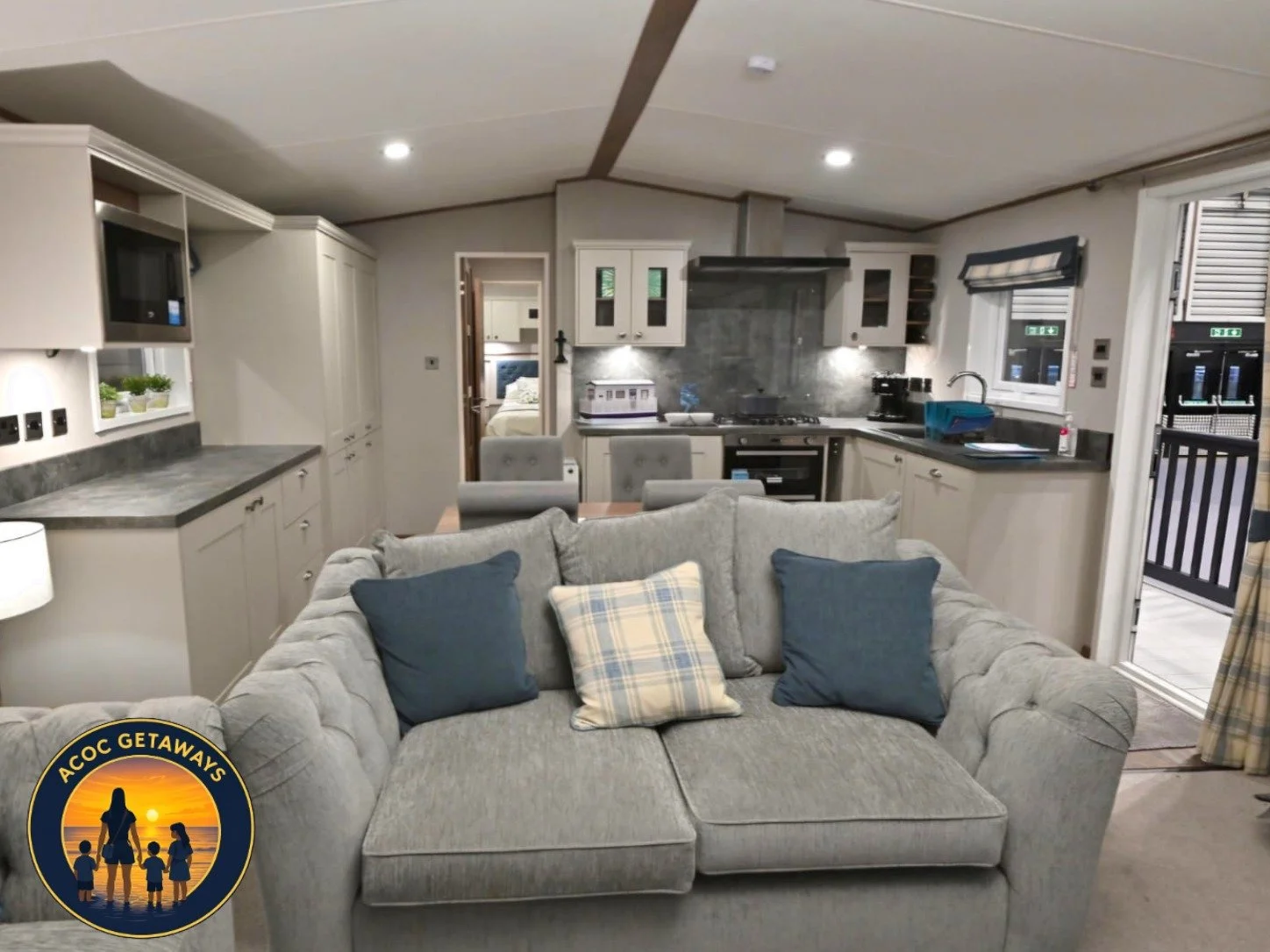 Living room and kitchen area of a cozy cabin or home with gray couch, blue and checkered pillows, kitchen cabinets, countertops, stove, and window with a view to a balcony.