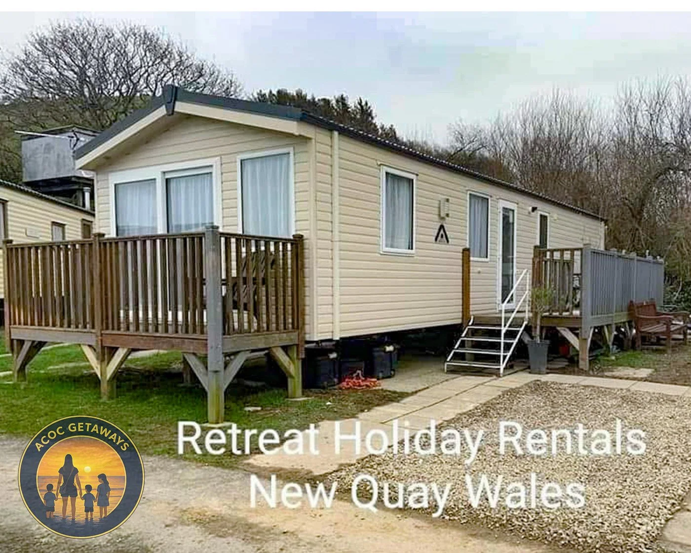 A beige mobile home with a raised wooden deck and metal stairs, situated on a grassy and gravel area, with leafless trees in the background, advertising retreat holiday rentals in New Quay, Wales, with a logo featuring a silhouette of a family at sunset.