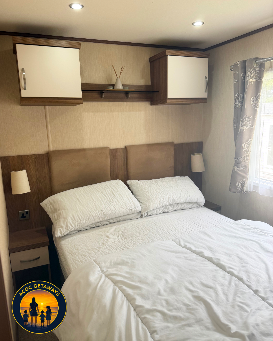 A cozy bedroom with a bed featuring a beige headboard and white bedding. Two bedside lamps are mounted on the wall beside the bed. In the corner, there is a window with patterned curtains. Above the bed, there are wall-mounted cabinets and a shelf wi