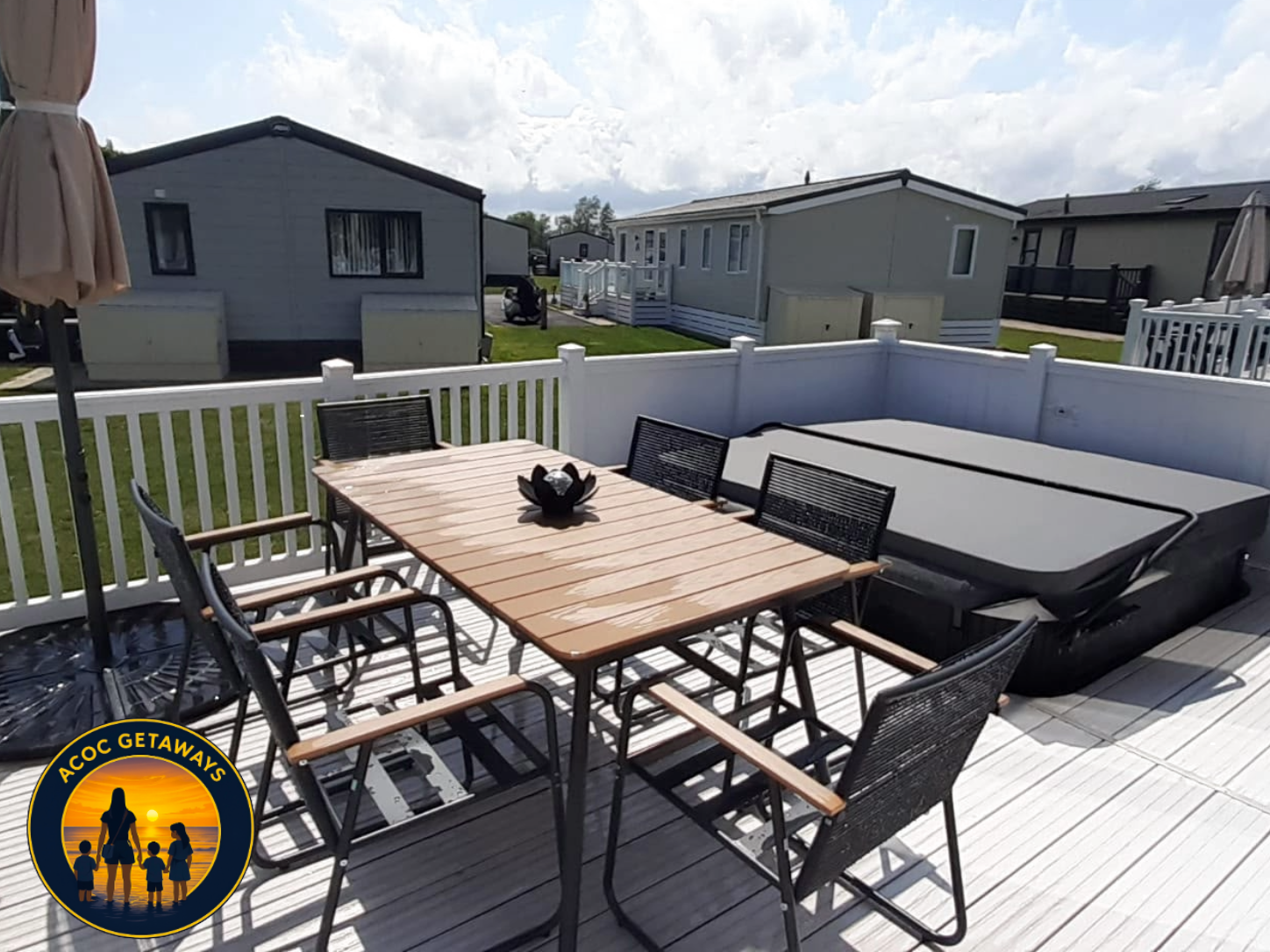 A backyard patio with a wooden dining table, six black chairs with armrests, and a hot tub enclosed by a white fence. There are umbrellas on the patio, and houses are visible in the background under a partly cloudy sky.