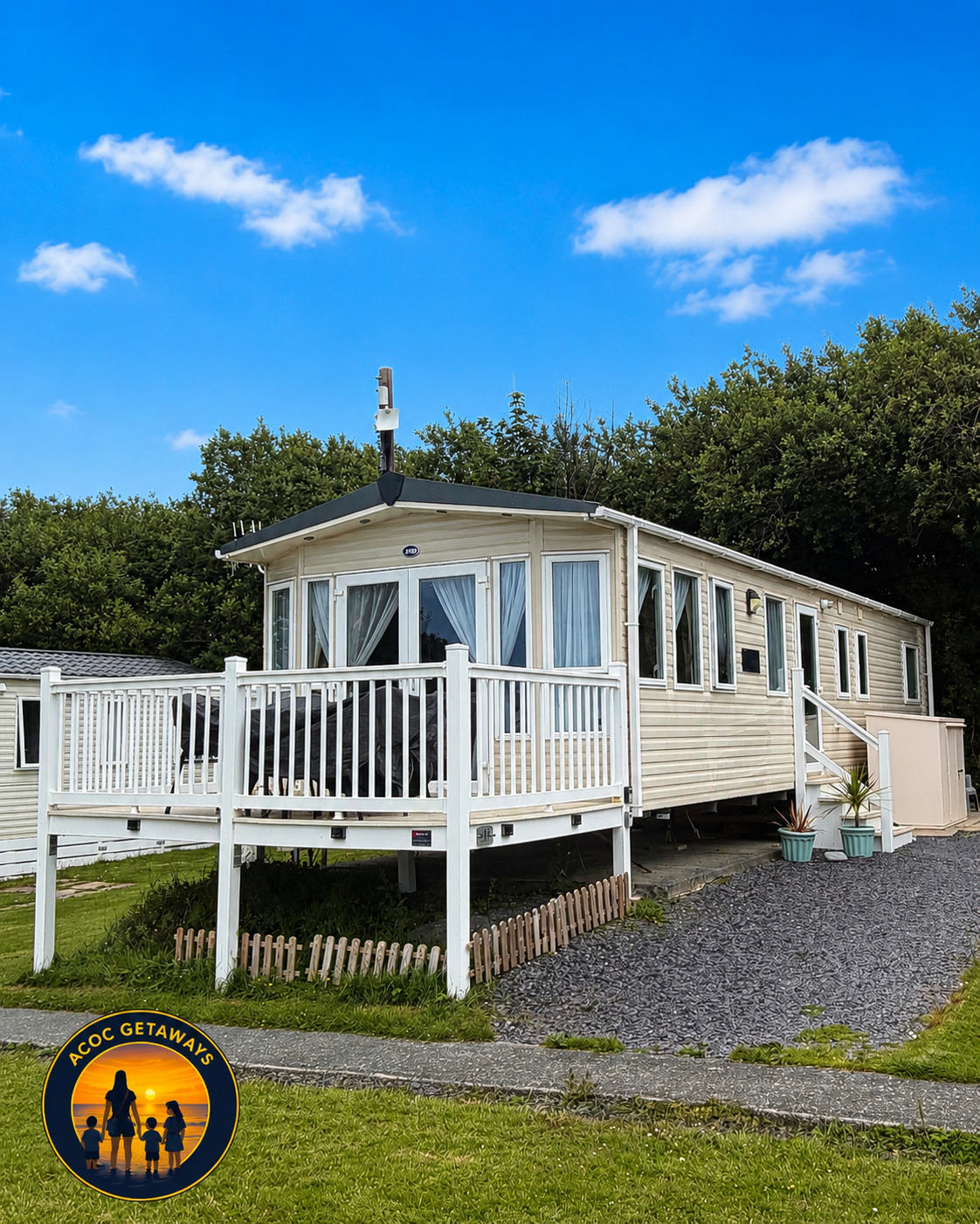 A white mobile home with a deck and railing, surrounded by greenery under a blue sky with clouds.