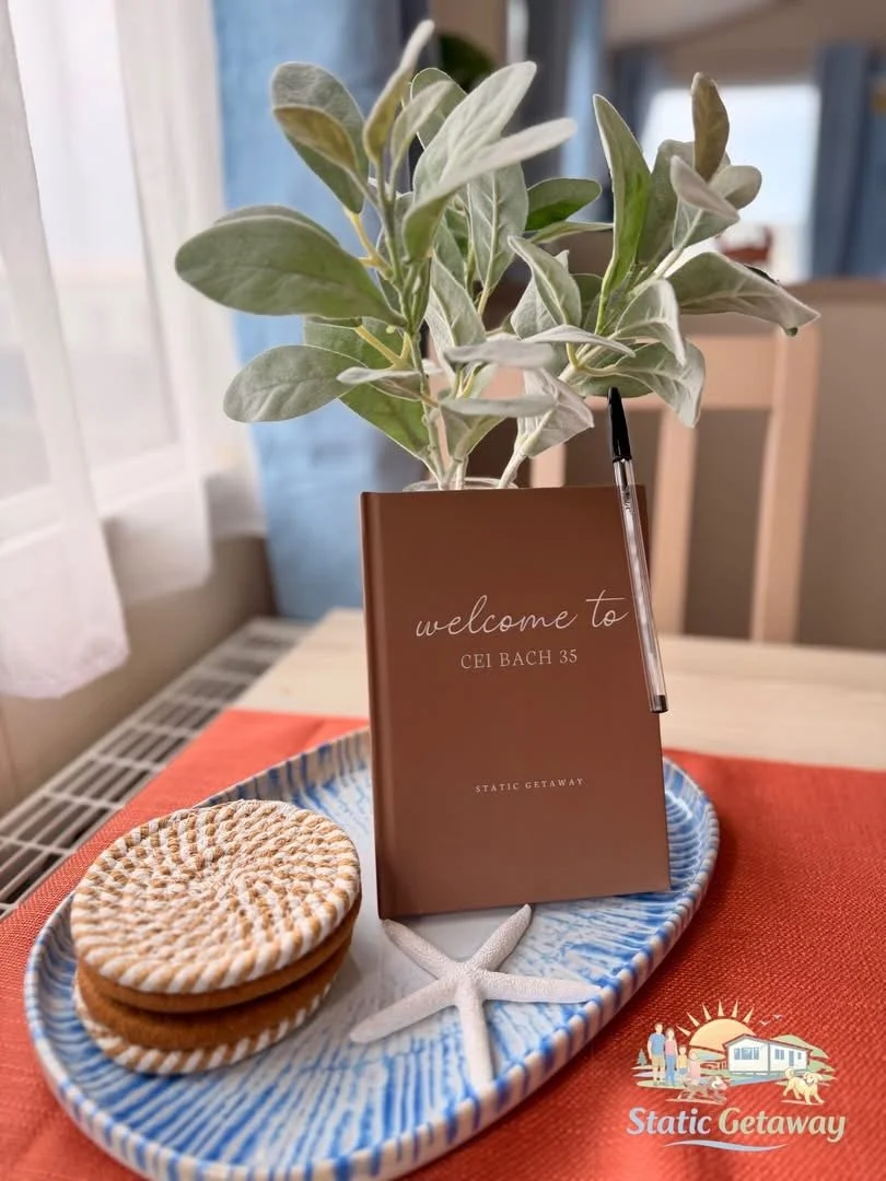 A decorative arrangement on a table, featuring a potted green and silver-leaved plant, a small brown welcome booklet titled 'Welcome to Cel Bach 35', a pen hanging from the booklet, two sandwich cookies with a crisscross pattern, a small white starfi