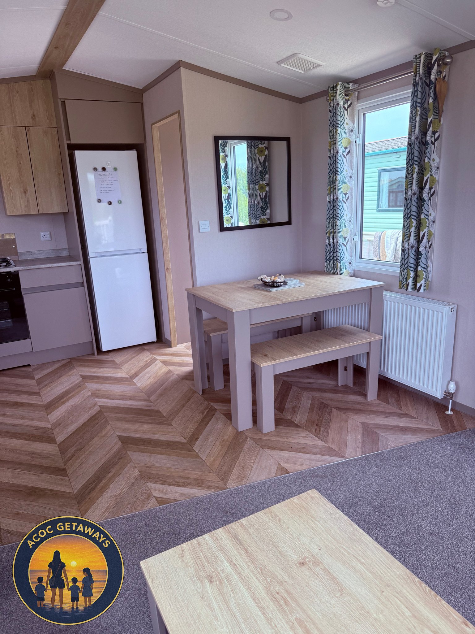 A compact kitchen and dining area with a wooden table and bench, a white frost-free refrigerator, patterned curtains, a wall mirror, a radiator, and a window showing neighboring house exterior.