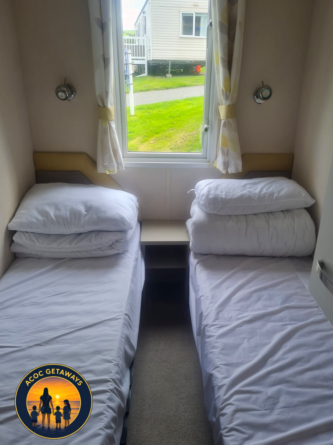 Two single beds with white bedding and pillows separated by a small nightstand in front of a window showing a grassy yard outside.