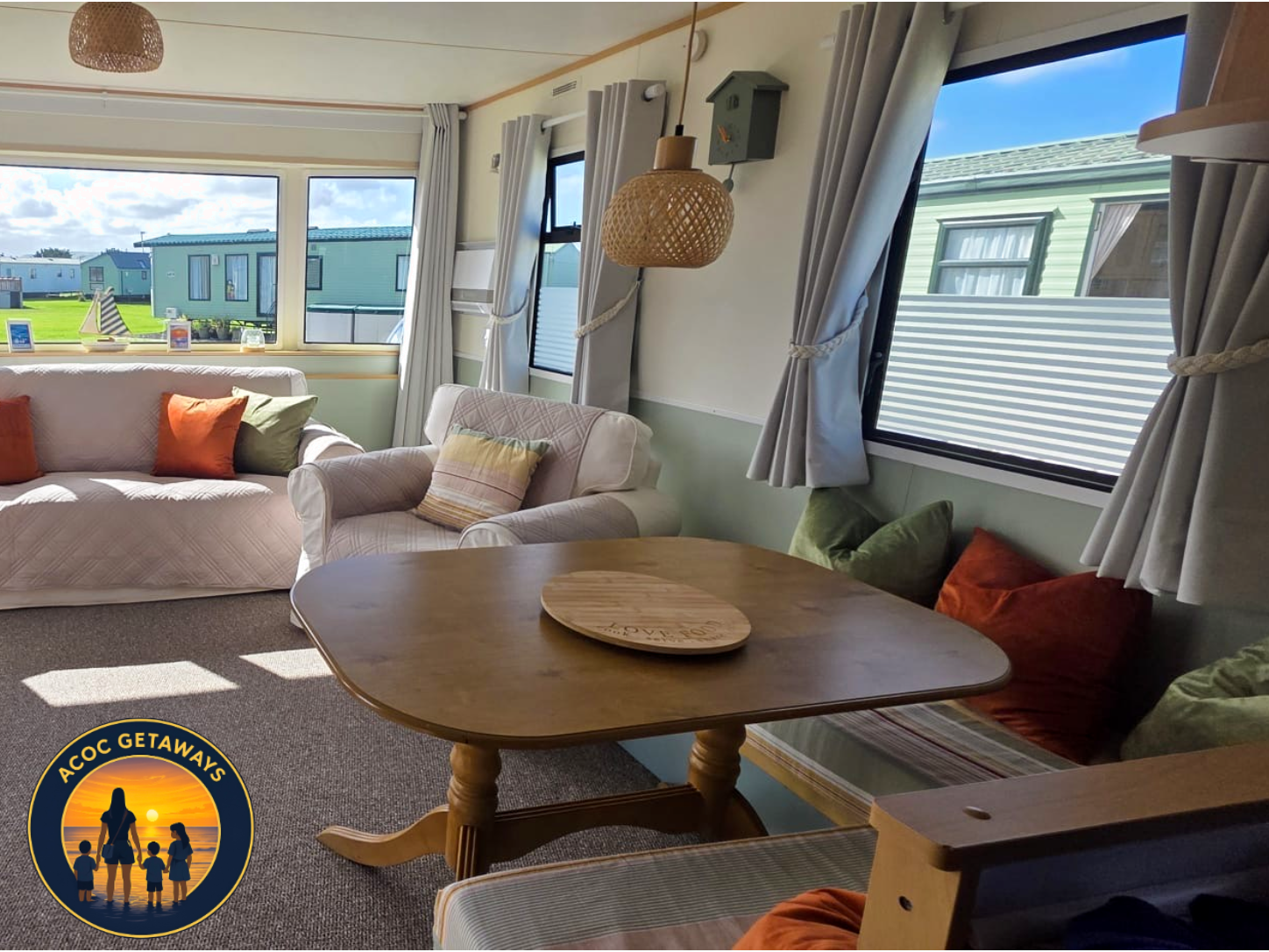 Living room with white sofa and armchair, colorful pillows, wooden table with a circular wooden tray, large windows with white curtains, and a view of green mobile homes outside, sunlight streaming in.