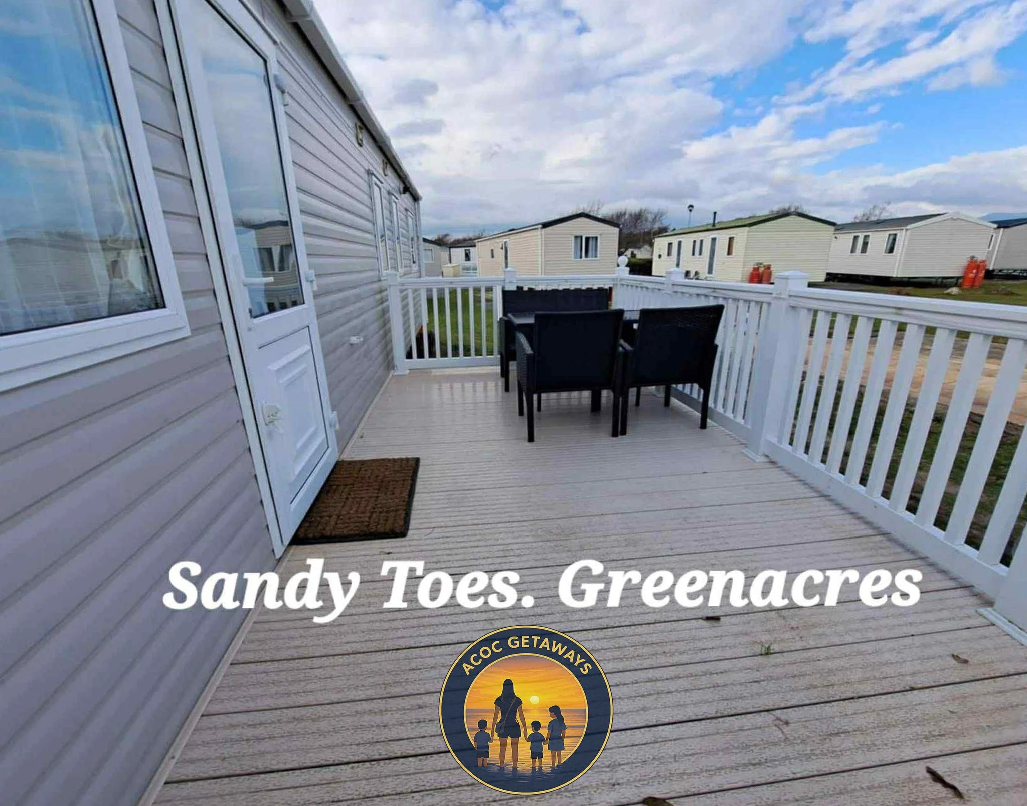 A spacious wooden deck with black outdoor table and four chairs, surrounded by a white railing, attached to a mobile home with beige siding, set in a community of similar mobile homes under a cloudy sky.