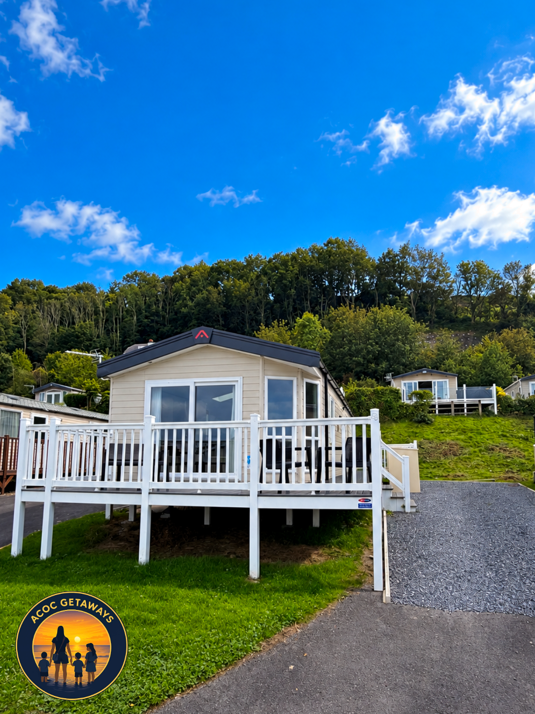 White house with a large deck elevated above ground, surrounded by green grass and trees, under a bright blue sky with scattered clouds.