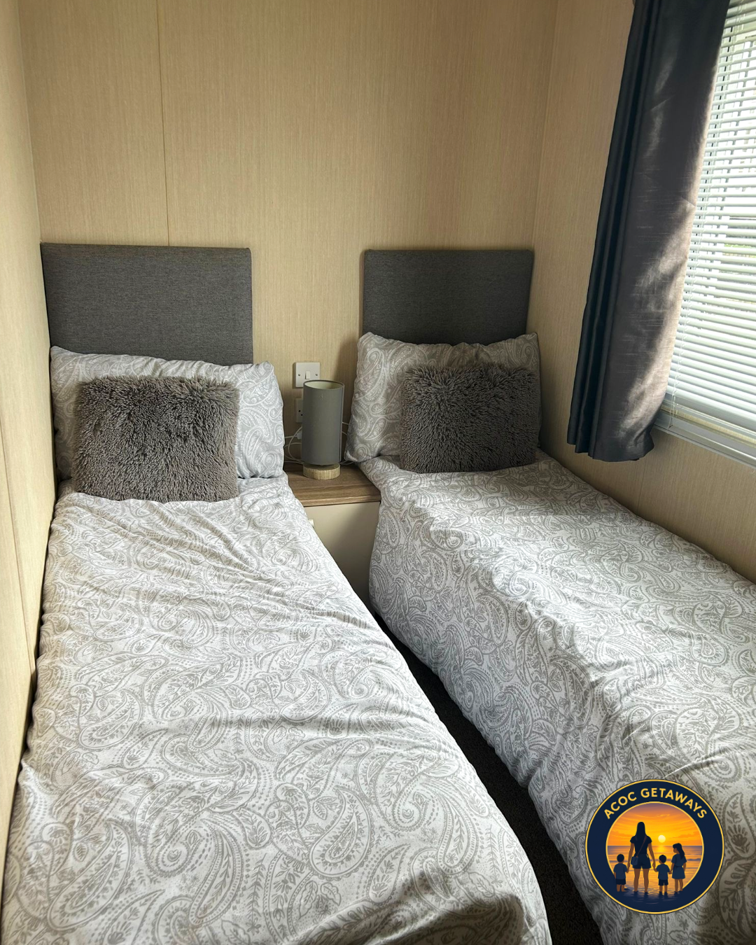 Two single beds with grey headboards, patterned white bedding, and grey throw pillows in a hotel room with a window and blinds. A nightstand with a lamp is between the beds.