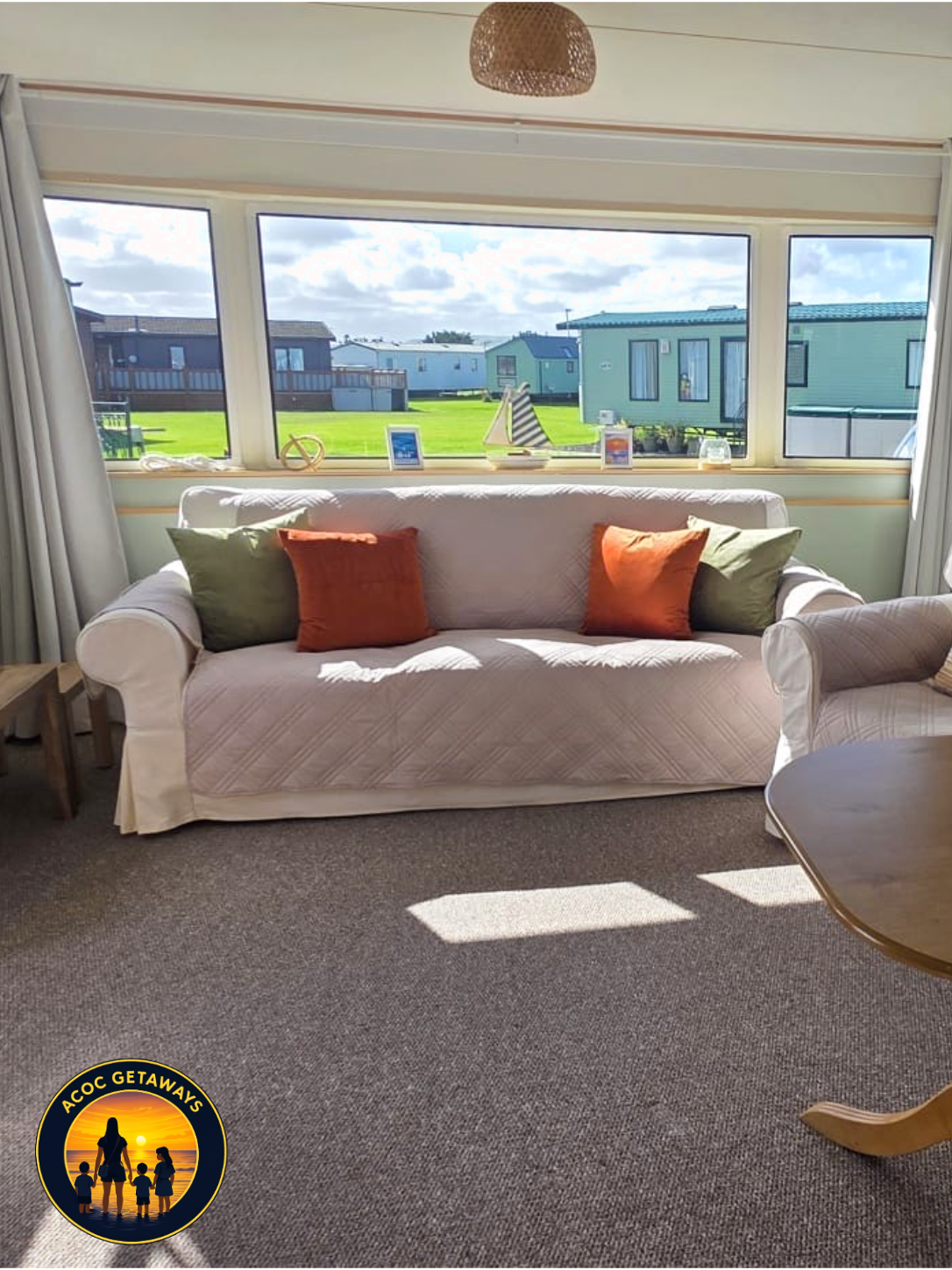 Living room with a white sofa, green and orange pillows, window view of green grass and buildings outside, small side table, and a wooden table partially visible on the right.