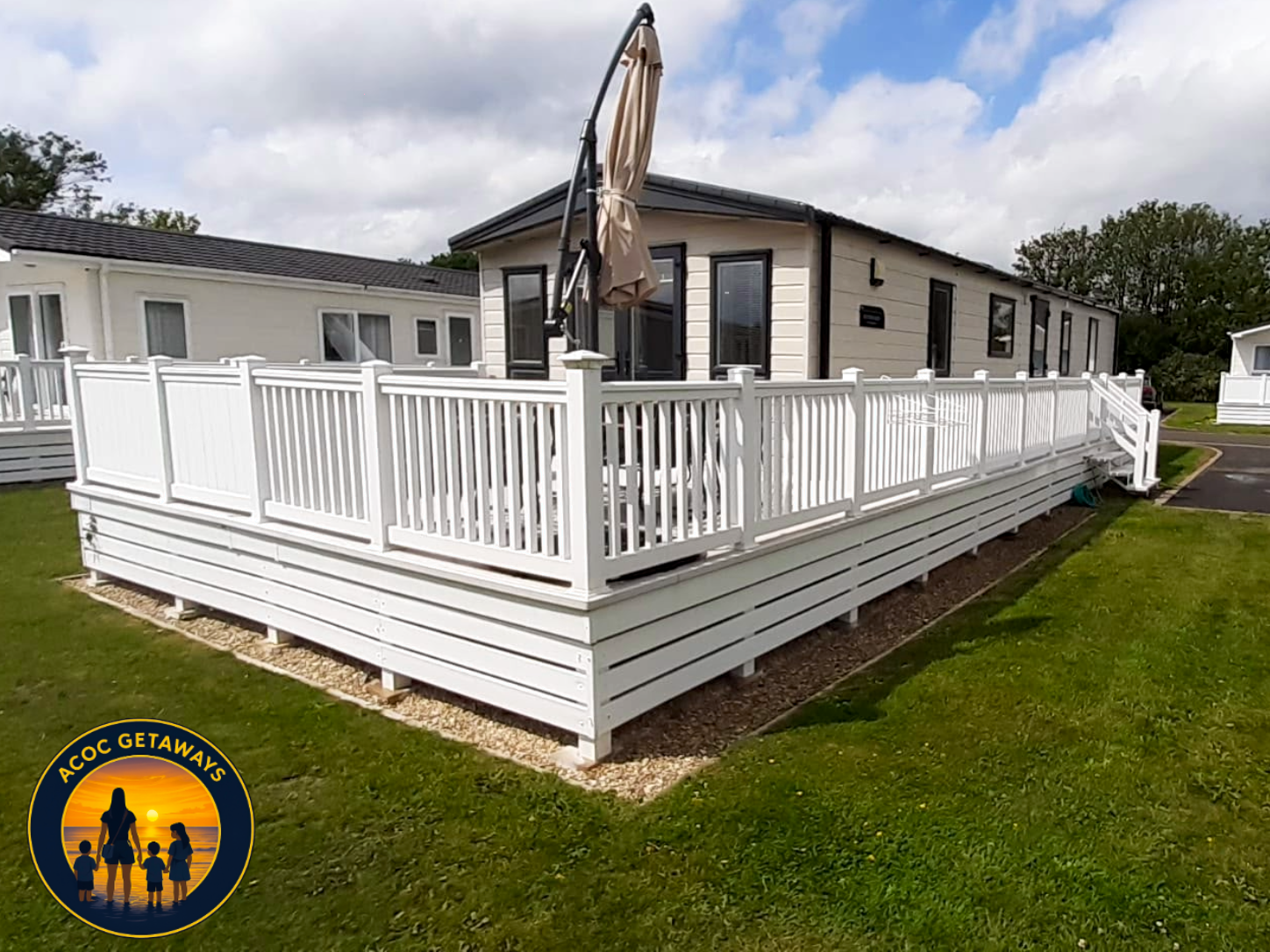 A white manufactured home on a raised foundation with a spacious porch enclosed by white railing, located on a green lawn under a partly cloudy sky.