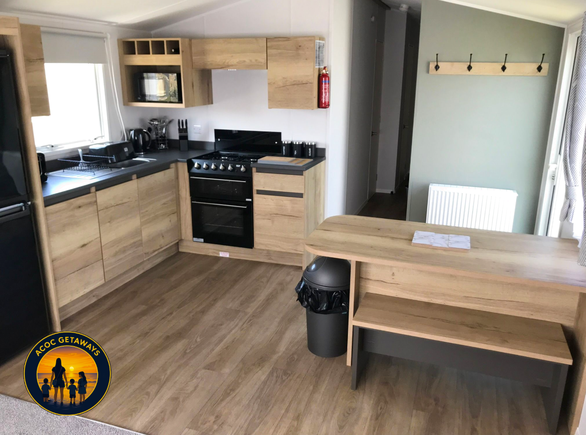 Kitchen with wooden cabinets, black appliances including oven and microwave, a black toaster, coffee maker, knife set, and window over the sink. There is a wooden breakfast bar with a trash can underneath, a small bench, a wall with hooks, and a fire