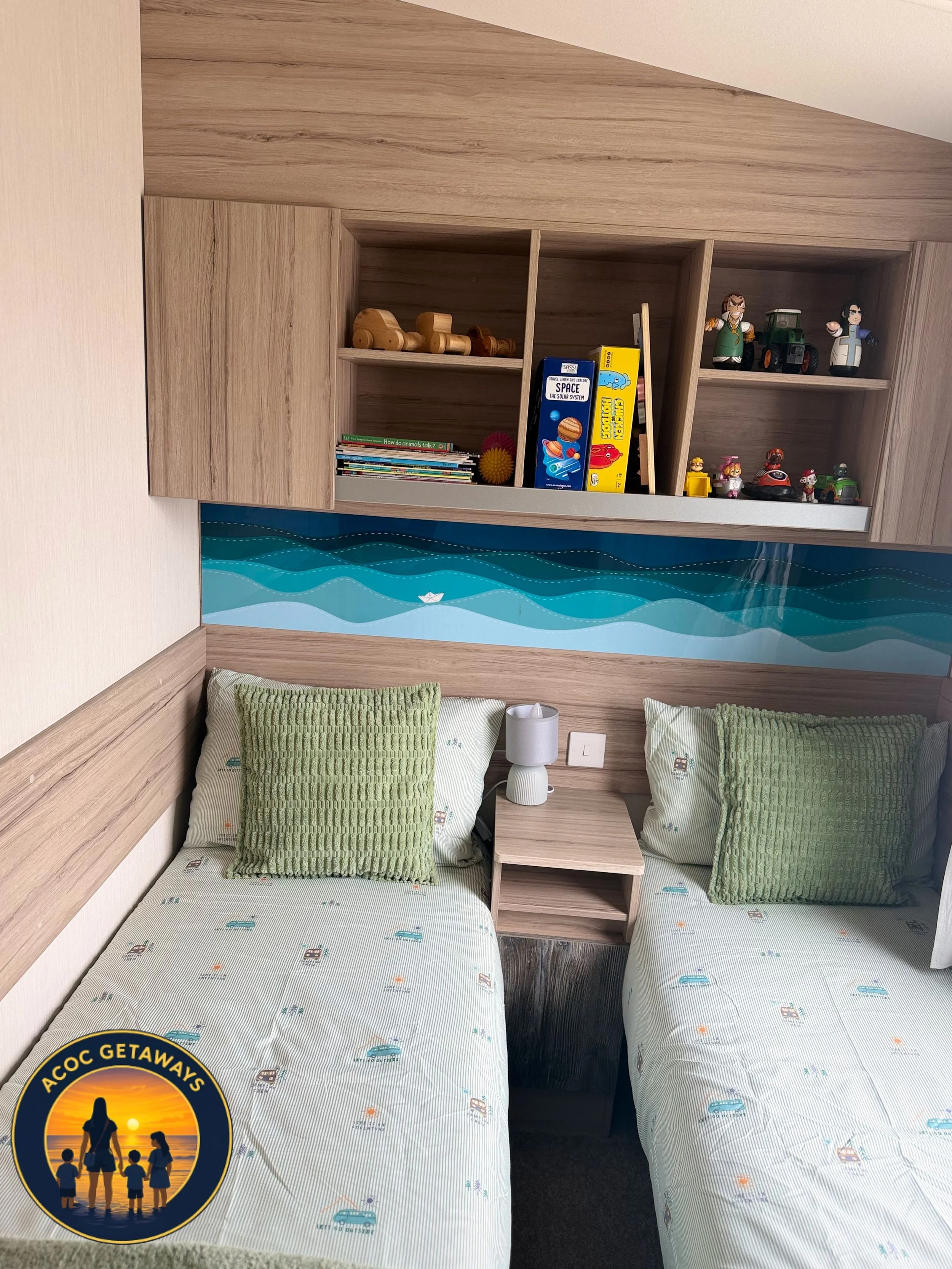 Two beds with light-colored sheets and green textured pillows, a small wooden nightstand with a lamp, a wooden shelf with books and toys mounted on a wood-paneled wall, blue wall with wave design, and an 'ACOC GETAWAYS' logo in the bottom left corner