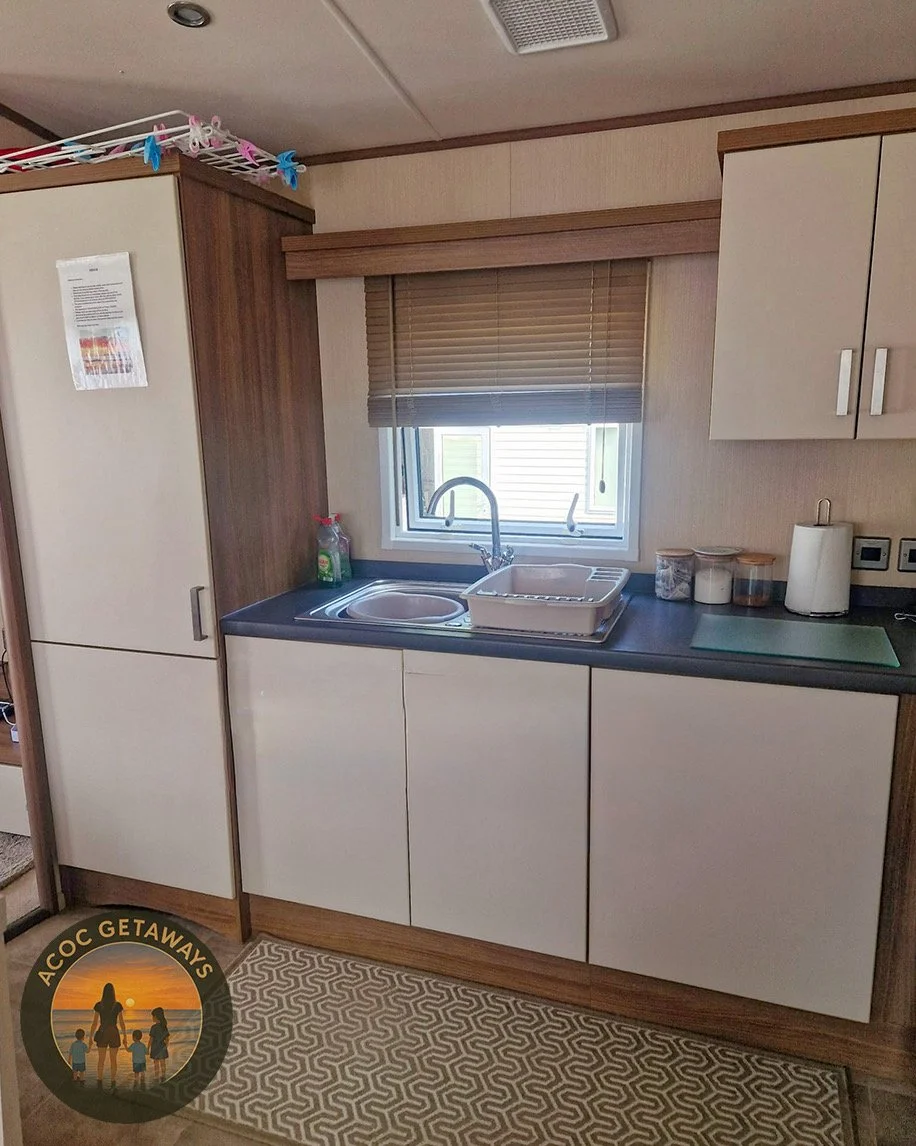 Small kitchen area with a window, sink, dish drying rack, and storage cabinets, next to a patterned rug on the floor.