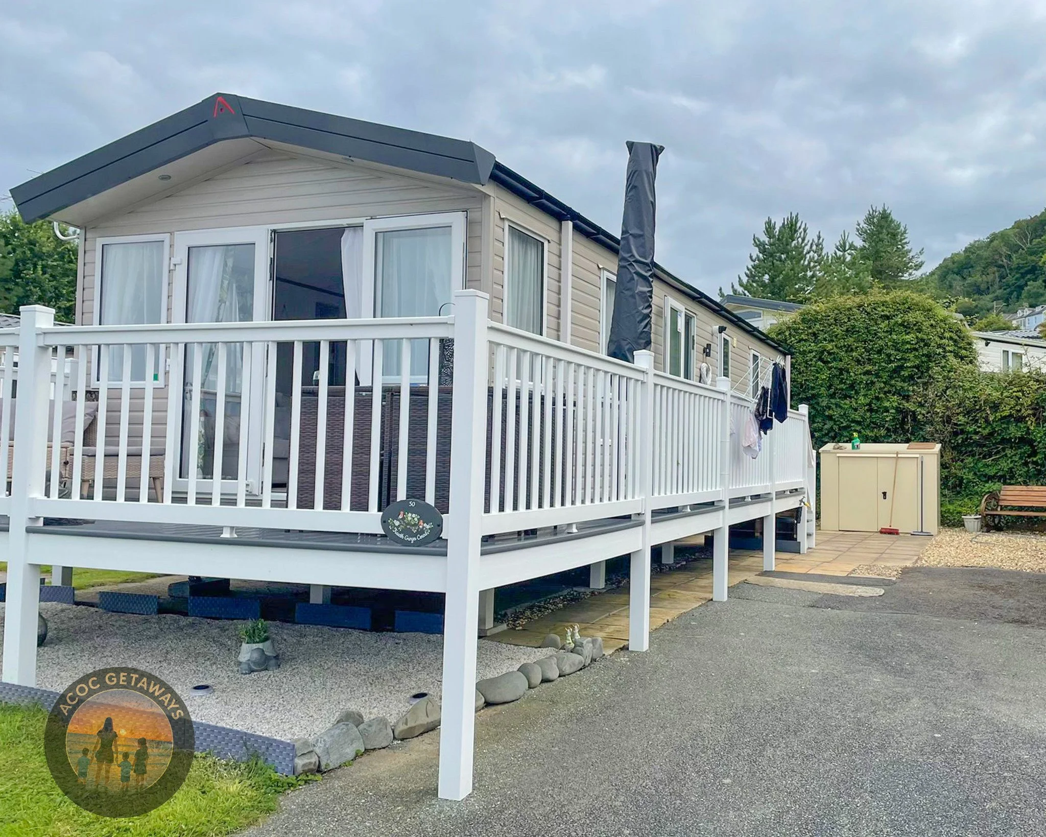 A mobile home with a white deck and railing, laundry hanging outside, and surrounded by trees and a driveway.