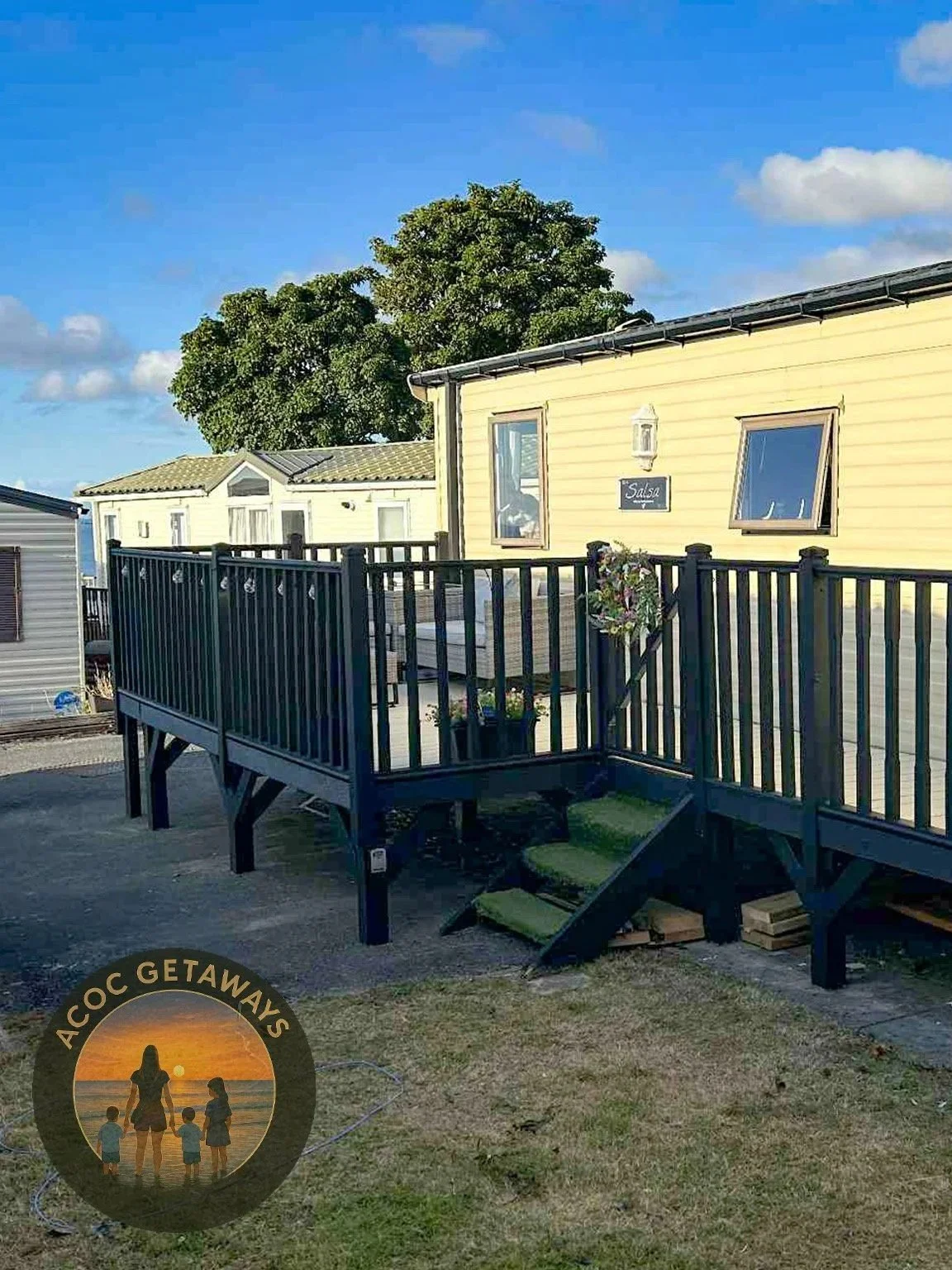 A yellow modular home with two tilted windows and a small outdoor deck with potted plants and stairs leading down to a grassy area. Clear blue sky with some clouds and a large green tree in the background.