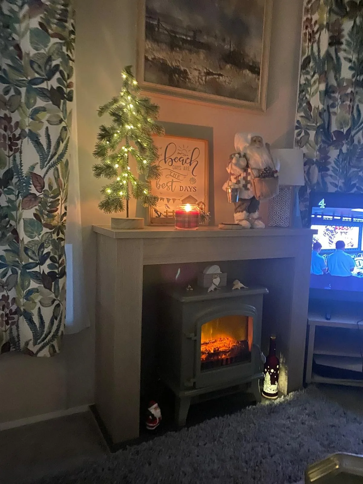 A cozy living room with a small decorated Christmas tree on the mantle, a Santa Claus figure, a lit candle, and a fireplace with a glowing fire, with a TV turned on to a game on the right.