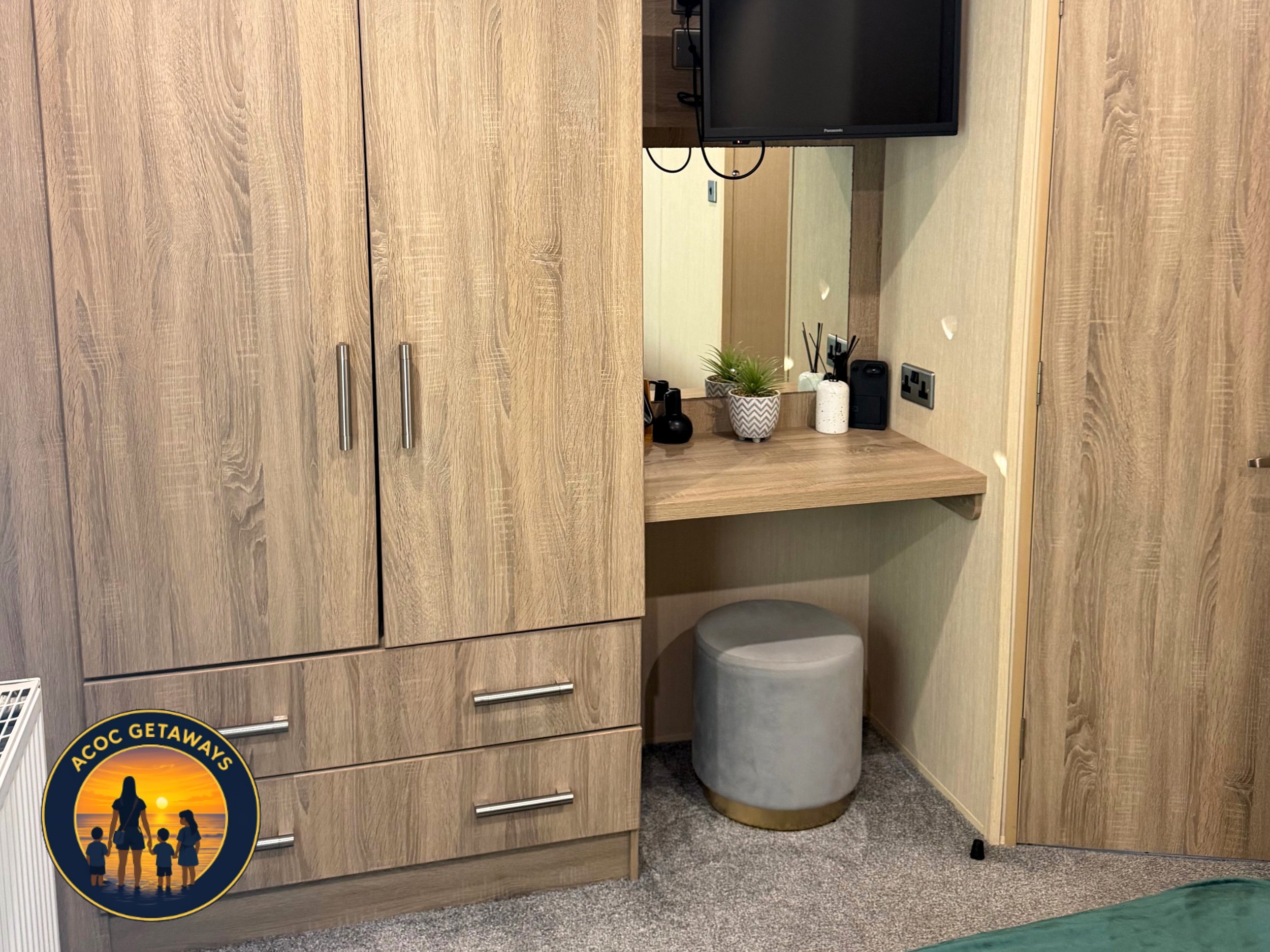 Wooden cabinet with silver handles, a small mirror, plants, candles, black decorative items, and a small gray ottoman, set against a wall with a black television and an electrical outlet.