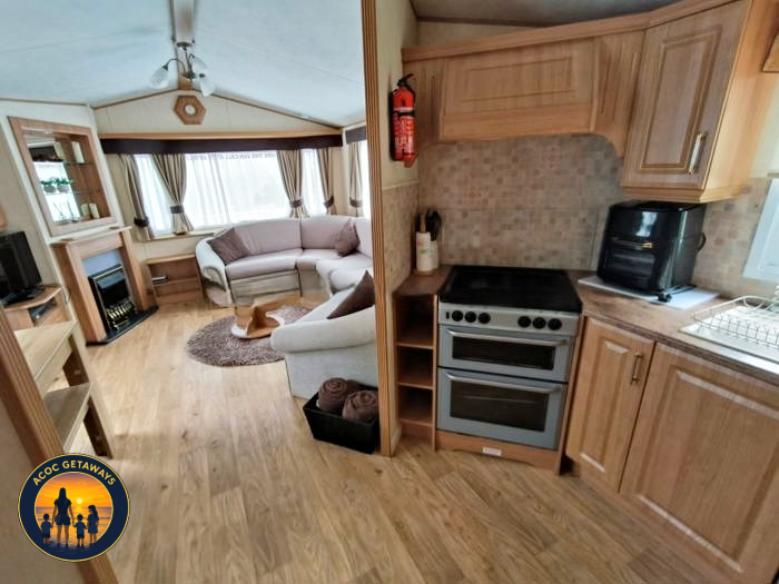 Living room with a beige couch, curved window with curtains, wooden furniture, and a fireplace; kitchen with wooden cabinets, stove, and small appliances.
