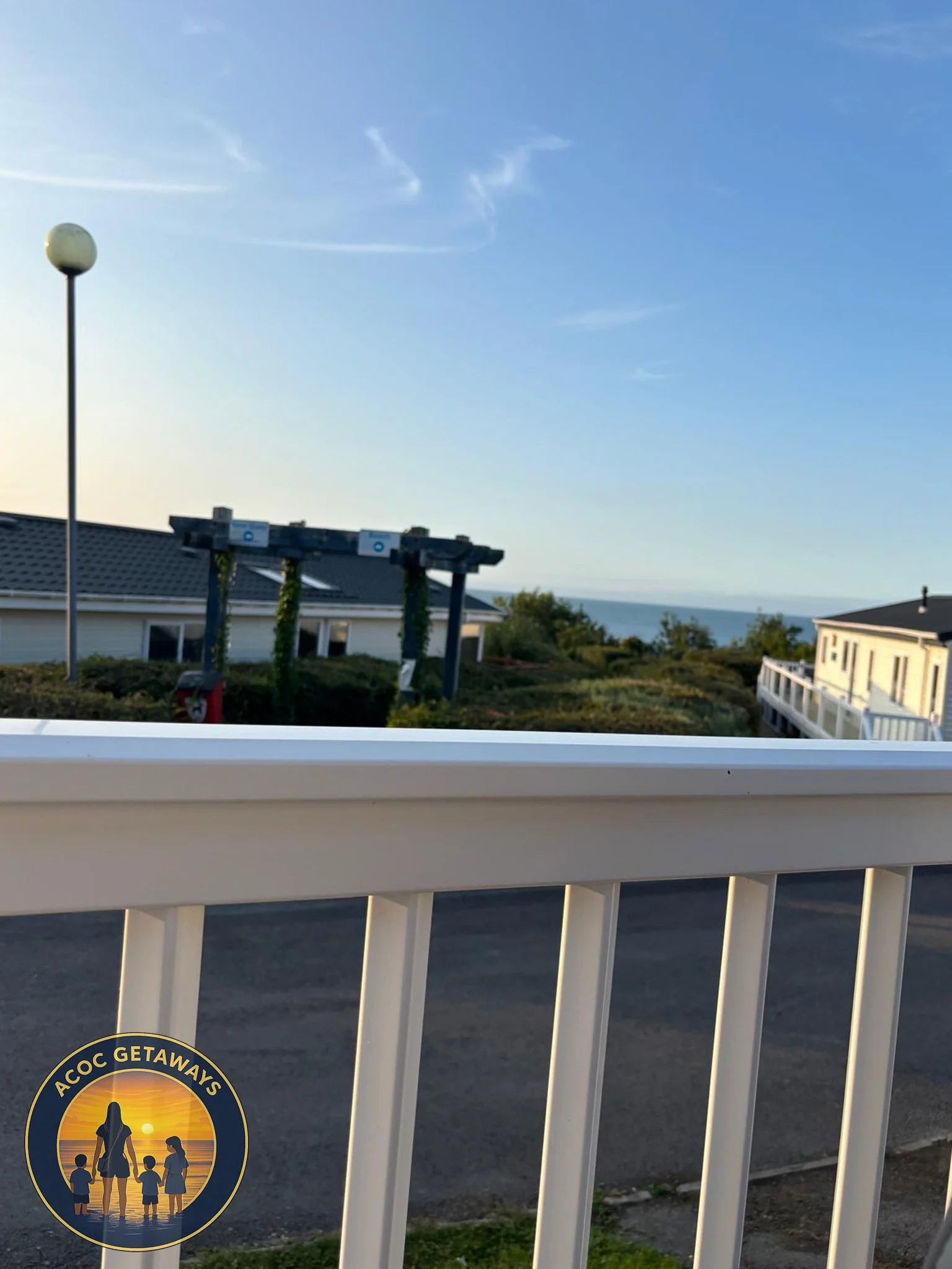 View of a suburban neighborhood overlooking the ocean, with a street, houses, a lamppost, and a pergola partly covered in greenery, during sunset or sunrise.