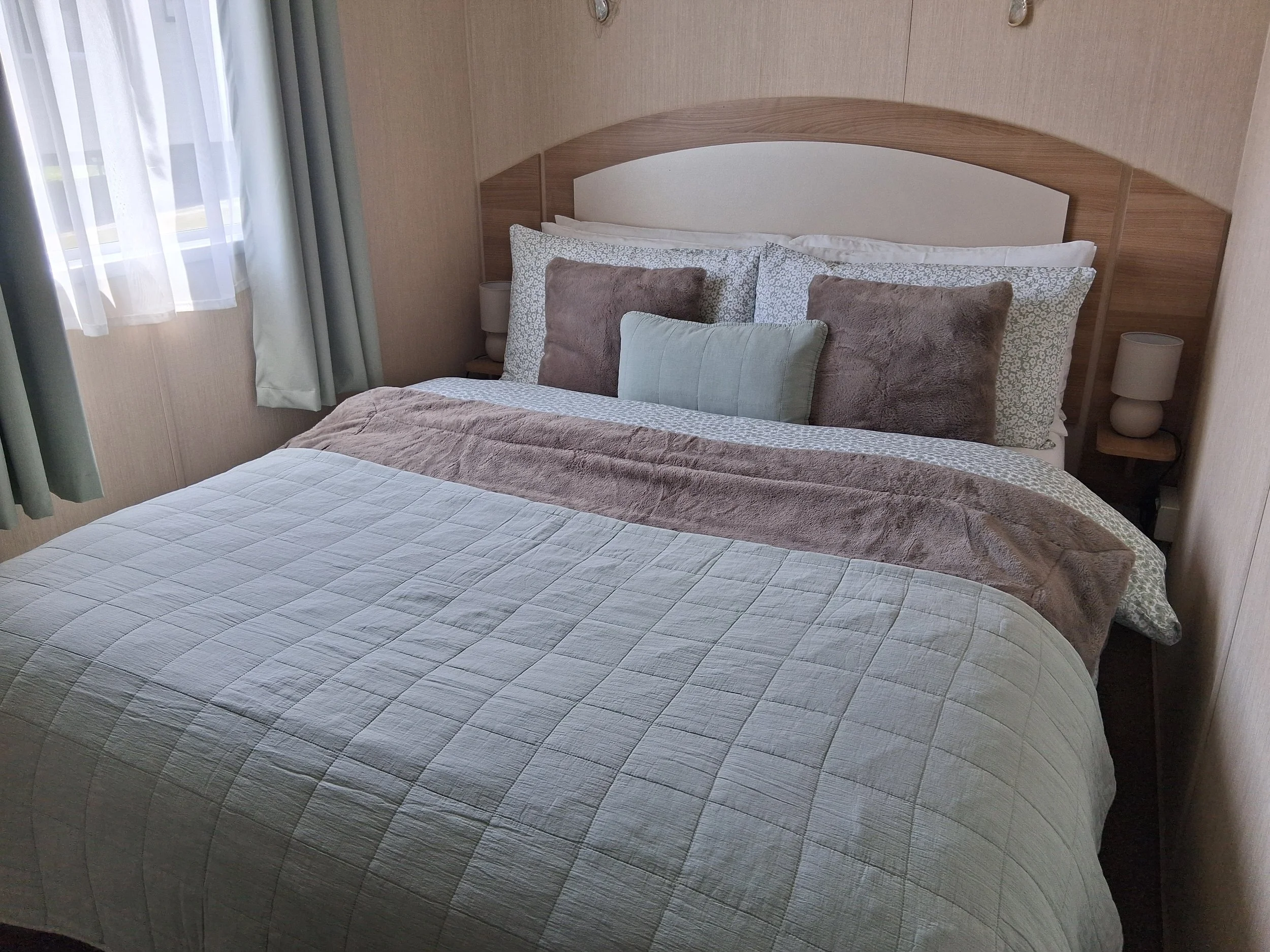 A neatly made bed with multiple pillows, a beige headboard, and two bedside lamps in a cozy bedroom setting, near a window with sheer curtains.