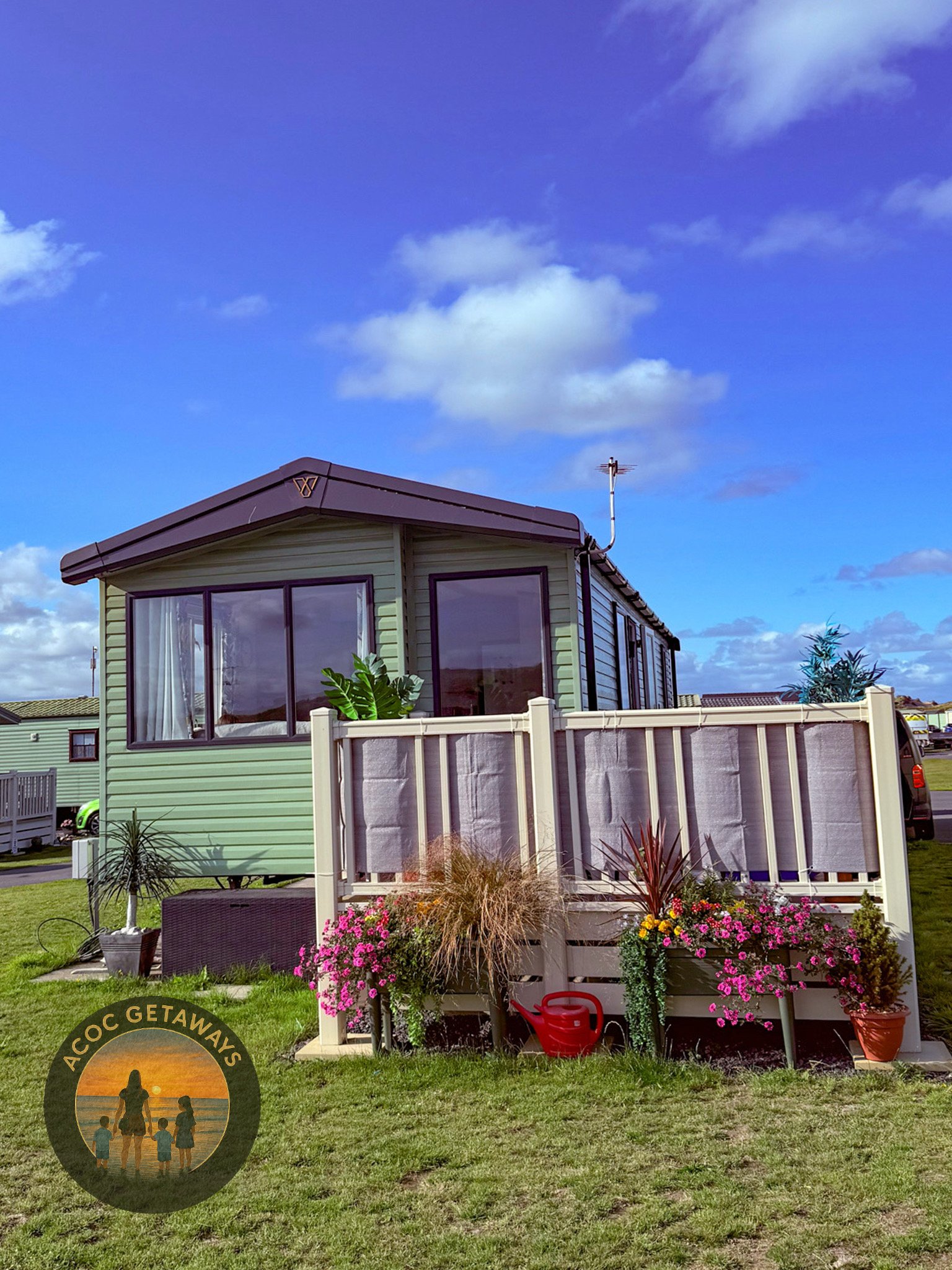 A small, pale green house with large windows, a deck with a white railing, potted plants, pink flowers, and a red watering can in a grassy yard under a partly cloudy blue sky.