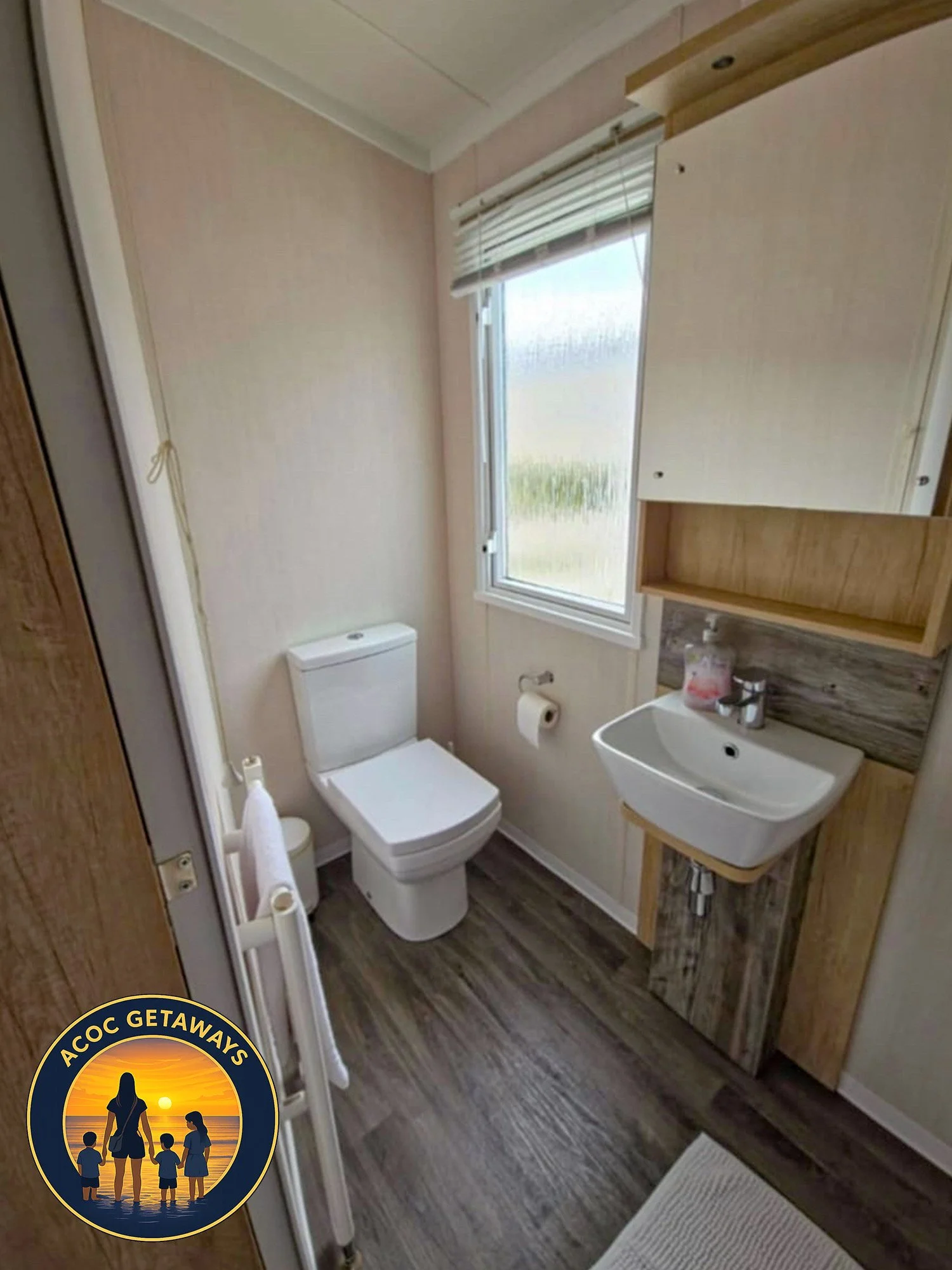 A small bathroom with a toilet, sink, and window, featuring wood and neutral-toned decor.