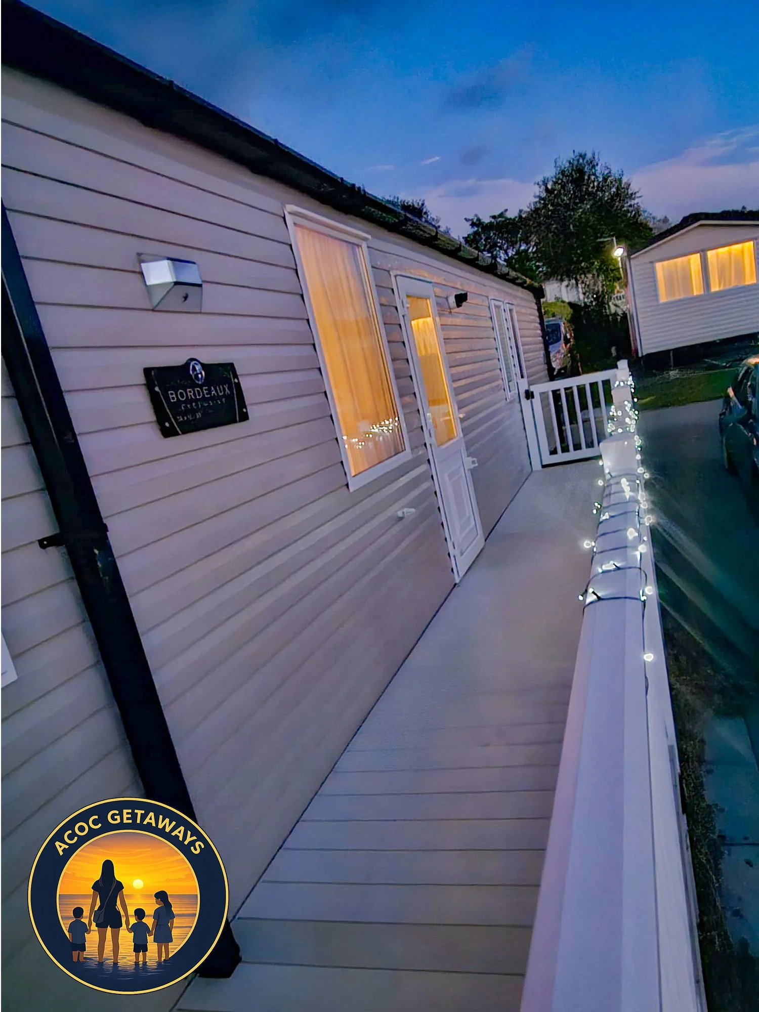 Exterior view of a mobile home with a porch decorated with string lights, at dusk, with another mobile home visible in the background, and a logo for ACCOC Getaways in the bottom left corner.